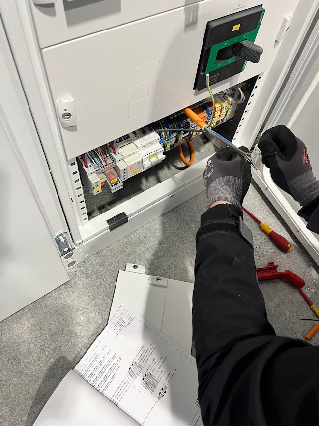 Elektryk w trakcie pracy nad rozdzielnicą elektryczną, używający narzędzi. Widoczne schematy, kable i zabezpieczenia. Oświetlenie sufitowe, szare tło podłogi.