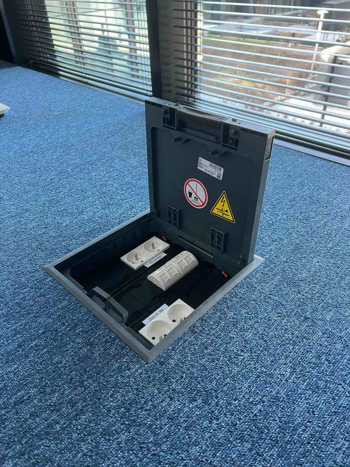 Otwarta podłogowa puszka elektryczna z gniazdami i złączkami na tle niebieskiej wykładziny dywanowej i okna z żaluzjami. Widoczne oznaczenia ostrzegawcze na pokrywie.