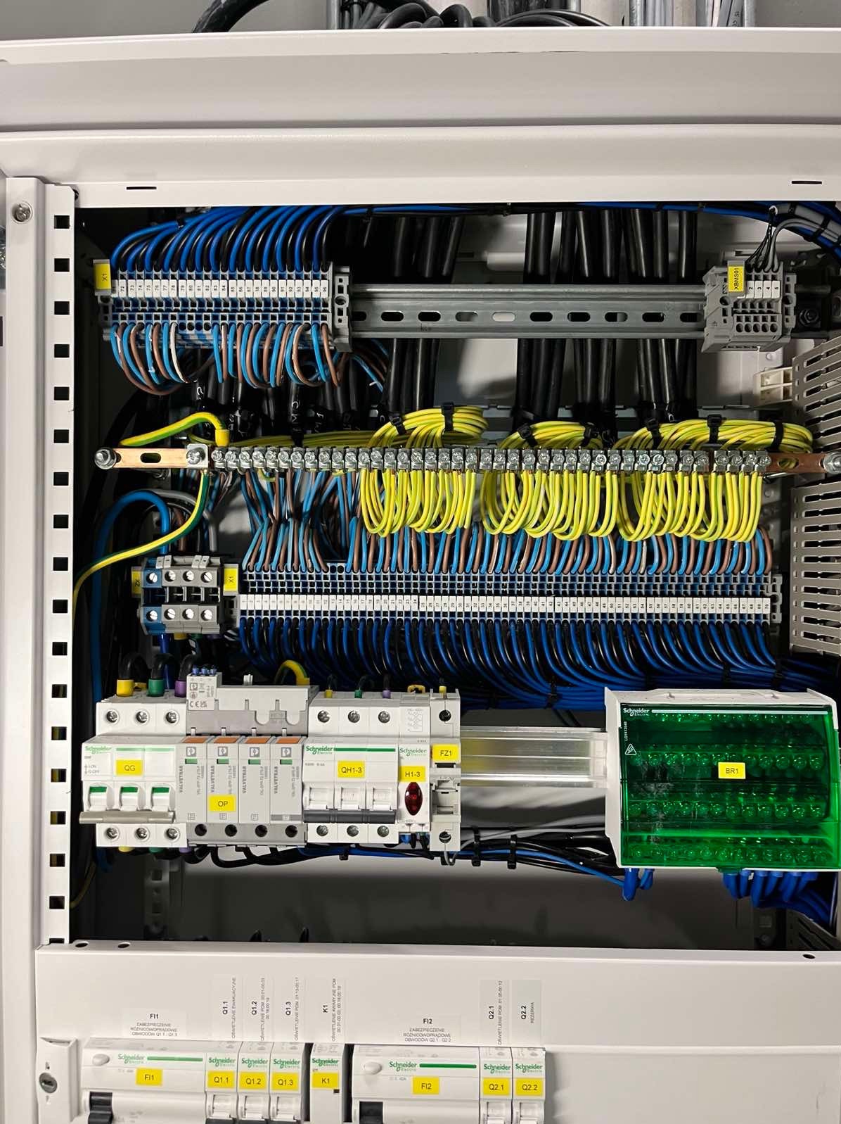 Rozdzielnica elektryczna z precyzyjnie ułożonymi przewodami w kolorach niebieskim i żółtym, zabezpieczenia Schneider Electric, widok z bliska na wnętrze szafy sterowniczej.