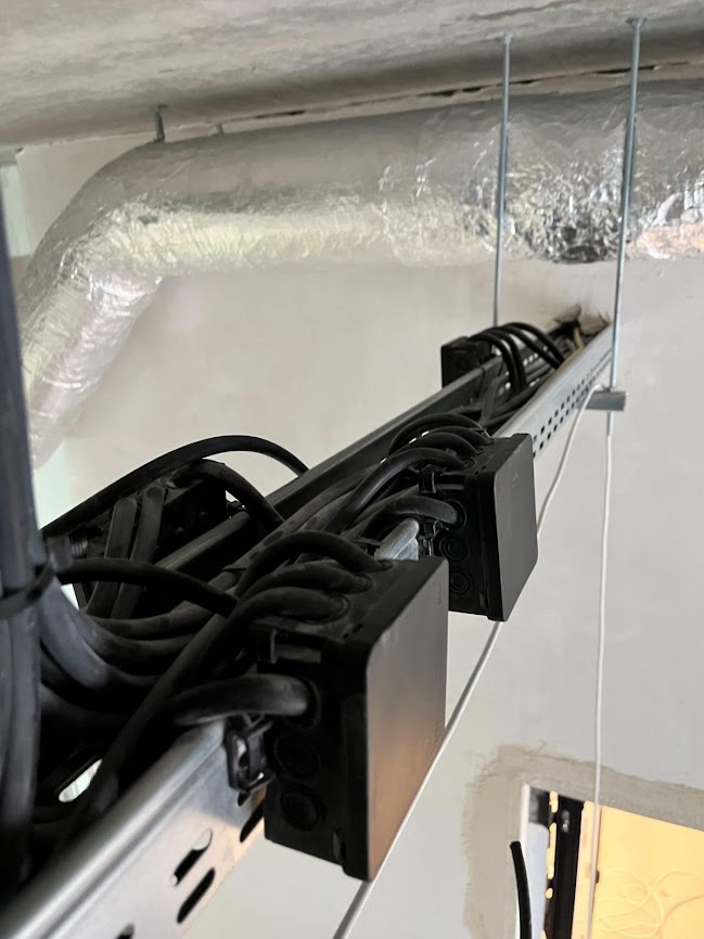 Metalowa szyna z czarnymi kablami elektrycznymi przymocowana do sufitu, widoczny fragment rury wentylacyjnej w izolacji. Ujęcie z dołu.