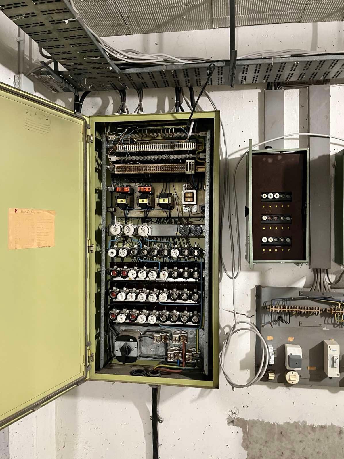 Otwarta, metalowa szafa elektryczna z bezpiecznikami i okablowaniem na ścianie w pomieszczeniu technicznym. Widoczne dodatkowe panele sterowania i kable w korytkach.