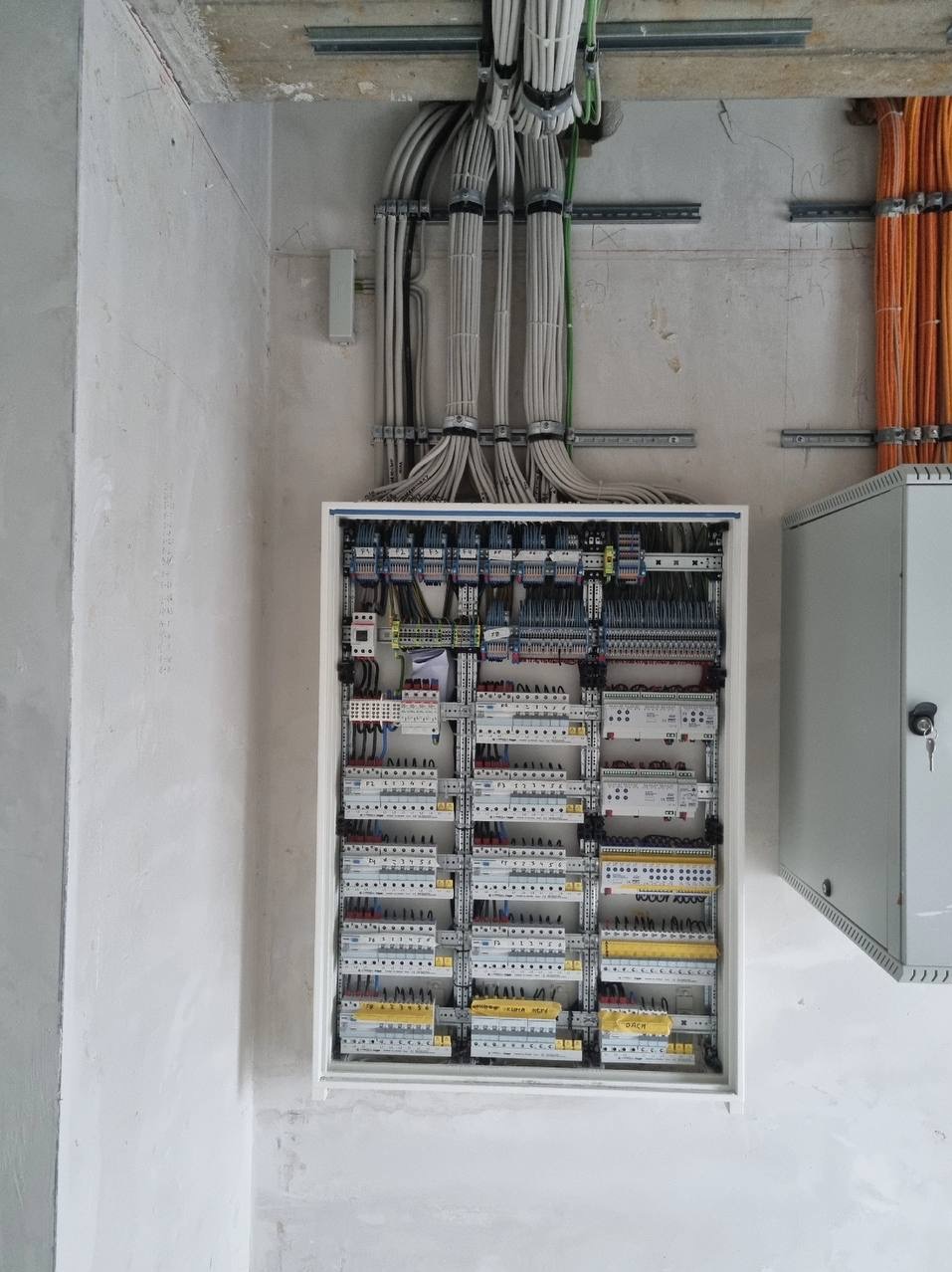 Rozdzielnica elektryczna z licznymi przewodami w nowym budynku w Warszawie. Widoczne zabezpieczenia i podłączenia. Szare ściany w tle.