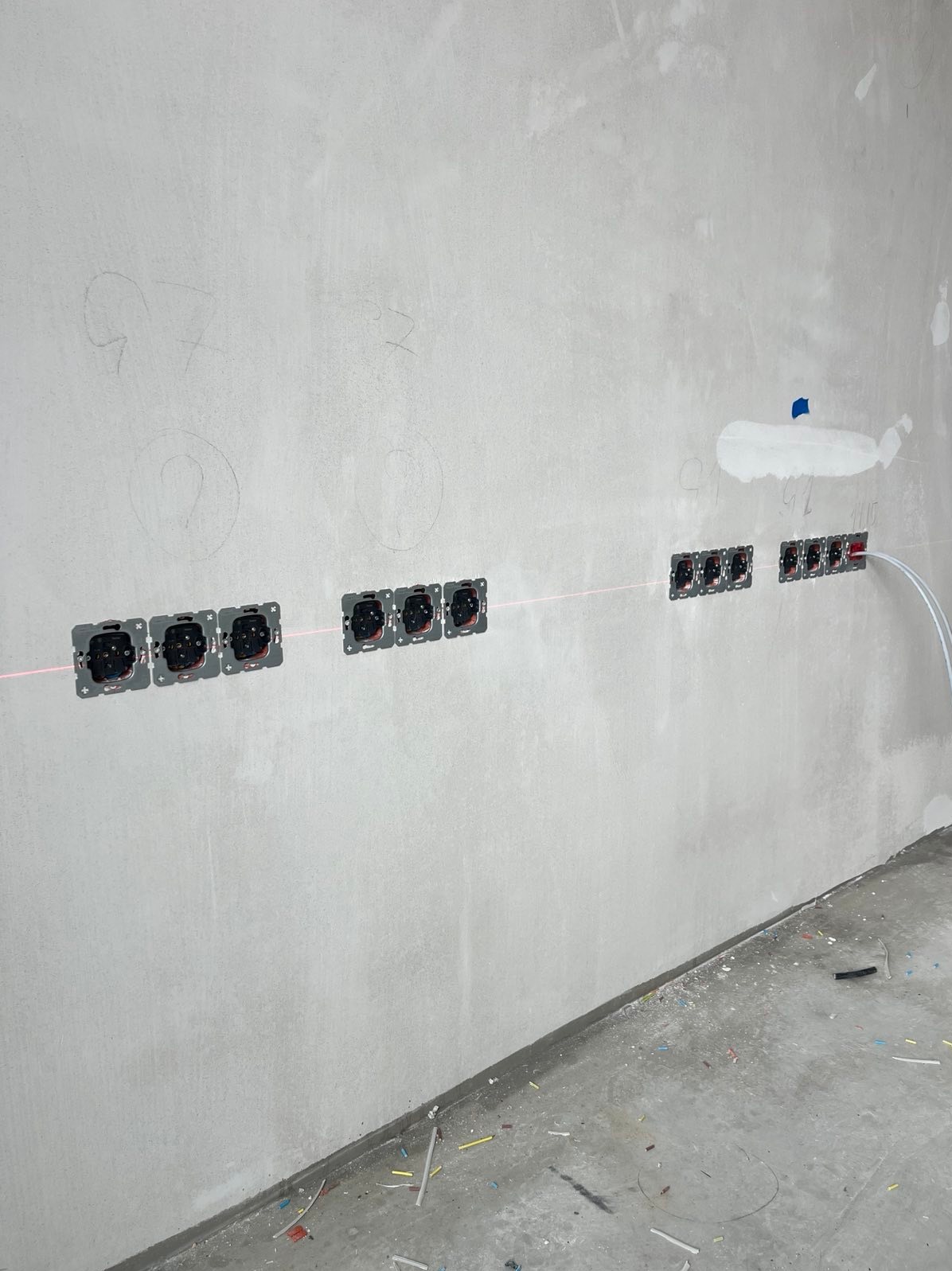 Szereg gniazdek elektrycznych w szarych ramkach, zamontowanych równo na surowej ścianie, wyznaczonych czerwoną linią lasera. Widoczne przewody.
