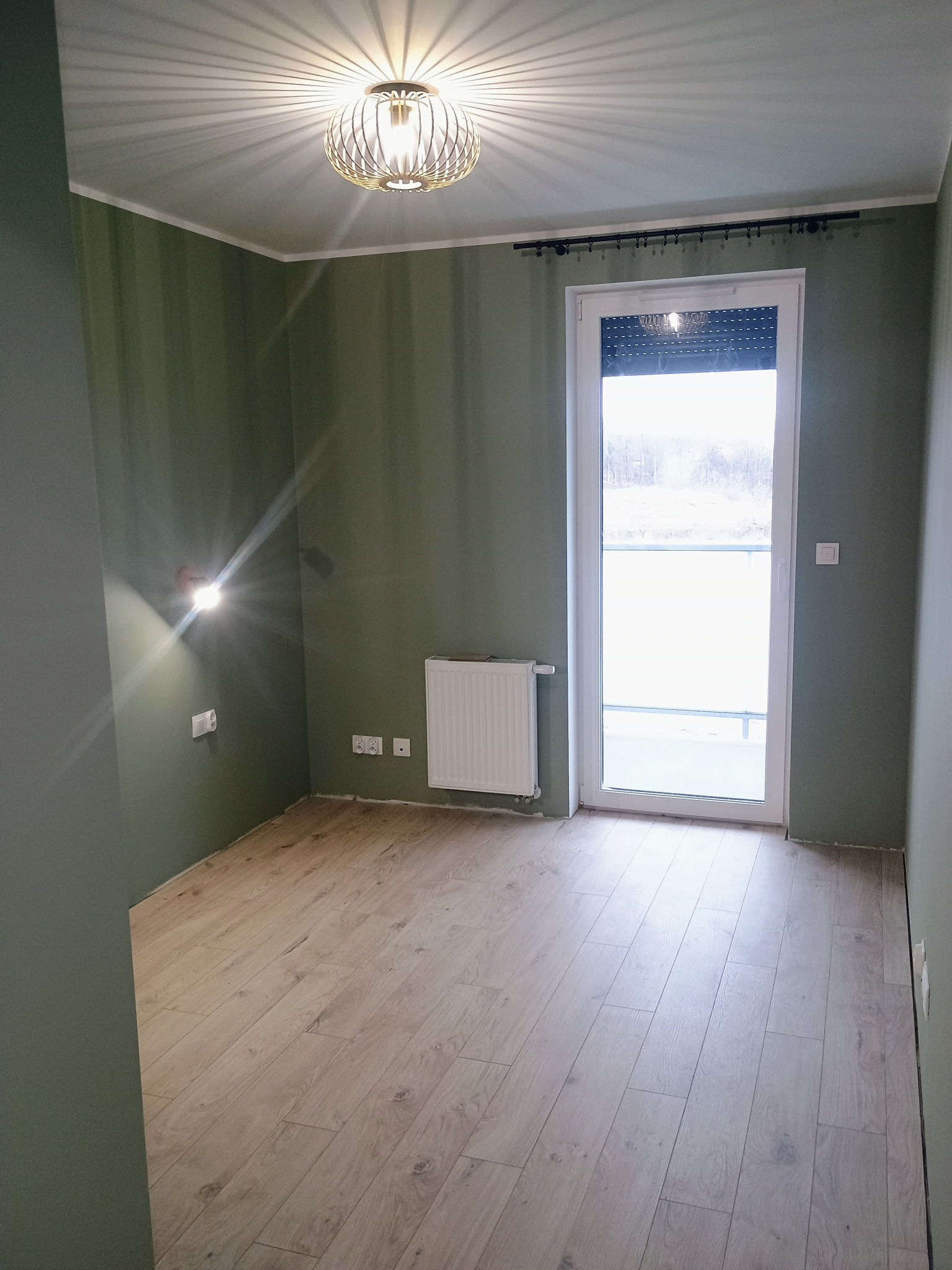 Pomalowane na oliwkowo ściany, jasna podłoga imitująca drewno i dekoracyjna lampa sufitowa w pustym pokoju po remoncie. Widoczne gniazdka elektryczne i grzejnik.