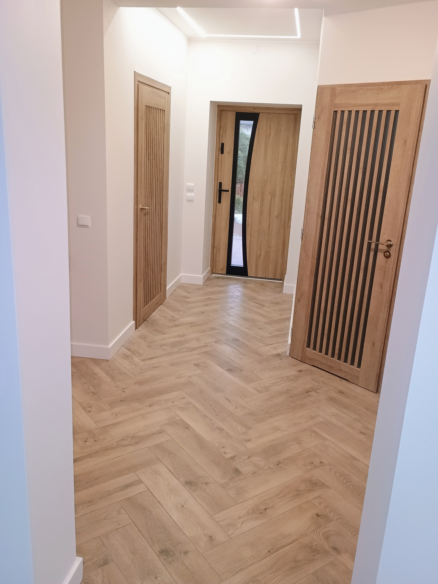 Jasny korytarz z parkietem w jodełkę, drewniane drzwi z pionowymi zdobieniami i białe ściany z listwami przypodłogowymi. Nowoczesne oświetlenie sufitowe.