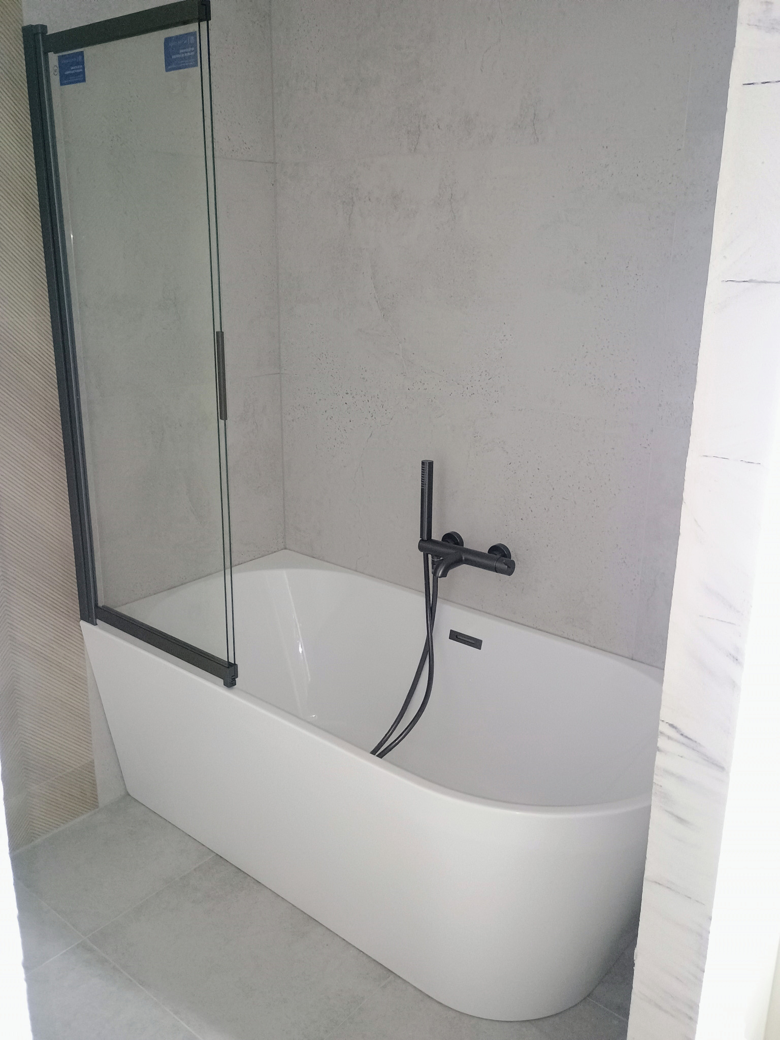 Nowoczesna łazienka: biała wanna z czarną armaturą i szklanym parawanem, szare płytki na ścianach i podłodze. Minimalistyczny design, jasne wnętrze.