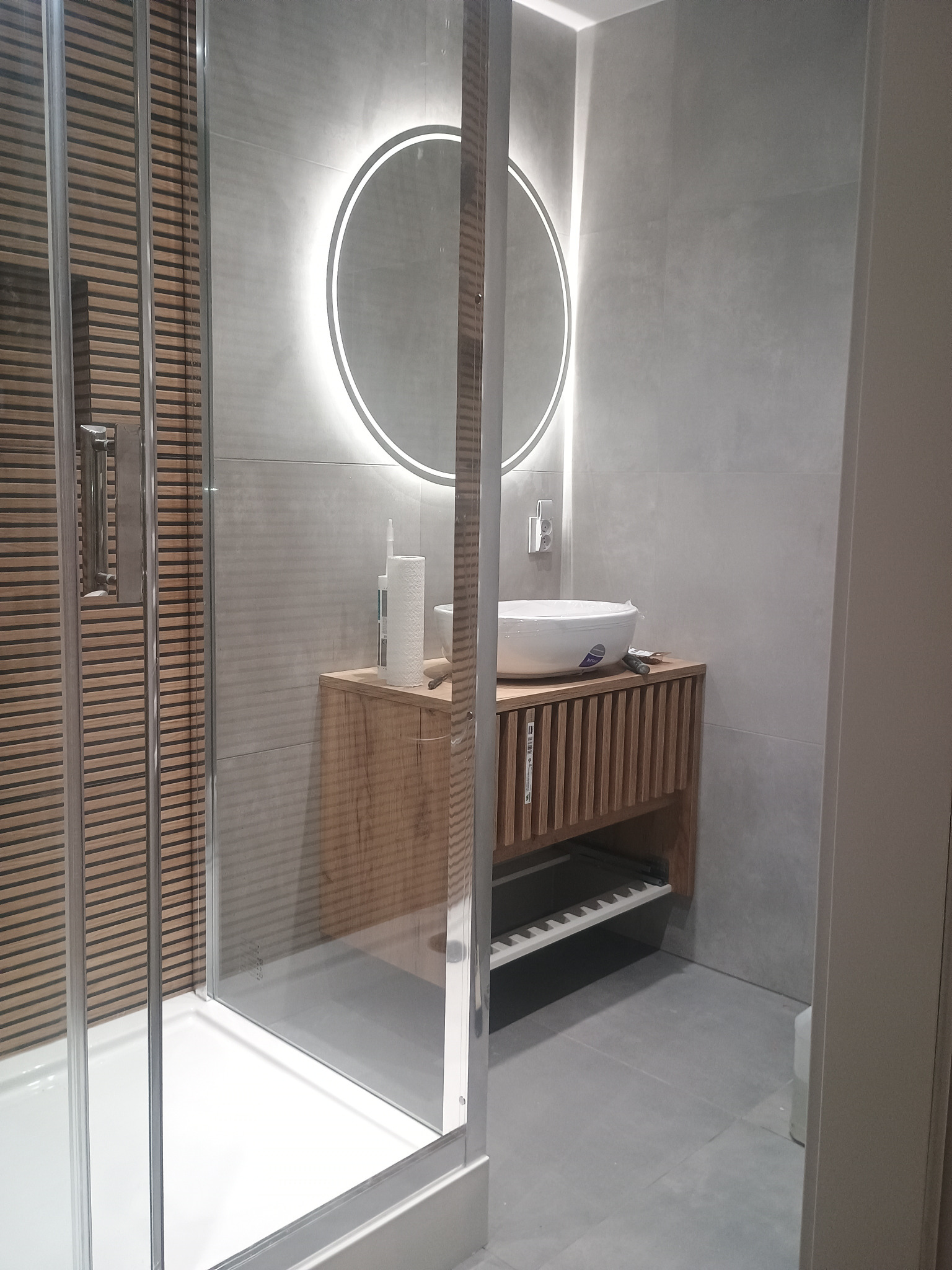 Nowoczesna łazienka z kabiną prysznicową, umywalką nablatową na drewnianej szafce i okrągłym lustrem z podświetleniem LED. Szare płytki na ścianach i podłodze.