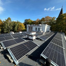 Vitalii Nykolyshyn - Instalacja paneli fotowoltaicznych na dachu budynku z widoczną wieżą kościoła w tle, jesienne drzewa i błękitne niebo. Częściowo widoczna osoba w odblaskowej kamizelce.