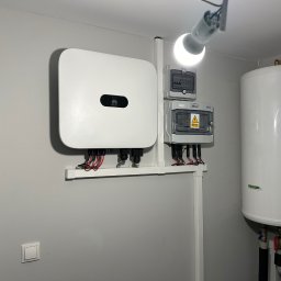 Vitalii Nykolyshyn - Biały inwerter Huawei i skrzynki elektryczne zamontowane na ścianie wewnątrz pomieszczenia, widoczne przewody i rury. Całość oświetlona żarówką.
