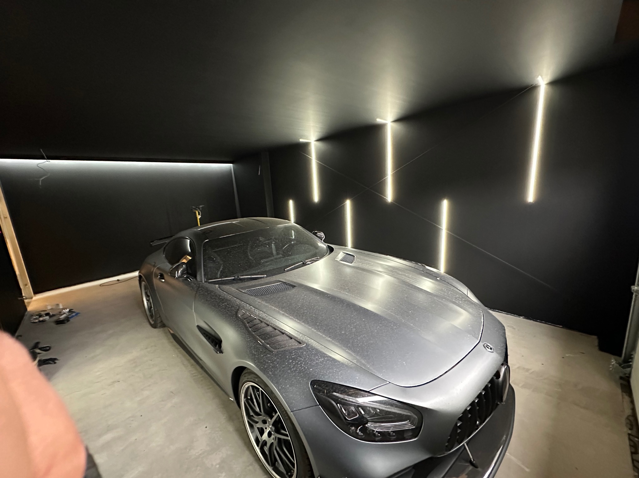 Szary sportowy samochód w garażu z nowoczesnym oświetleniem LED na czarnych ścianach, tworzącym geometryczny wzór.