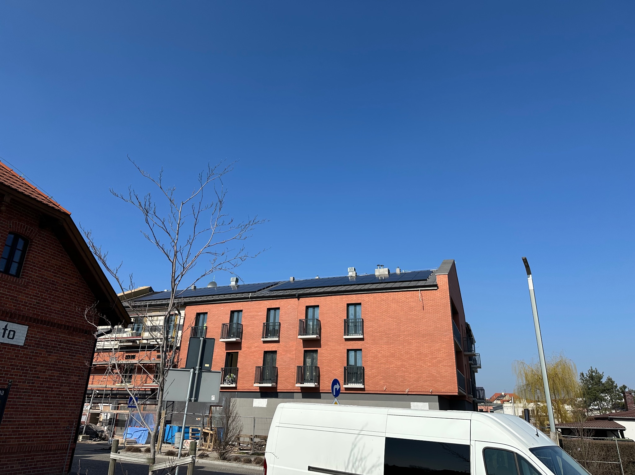 Ceglany budynek wielorodzinny z panelami słonecznymi na dachu, widoczny z poziomu ulicy, z białym vanem w kadrze. Częściowo widoczne rusztowania przy sąsiednim budynku.