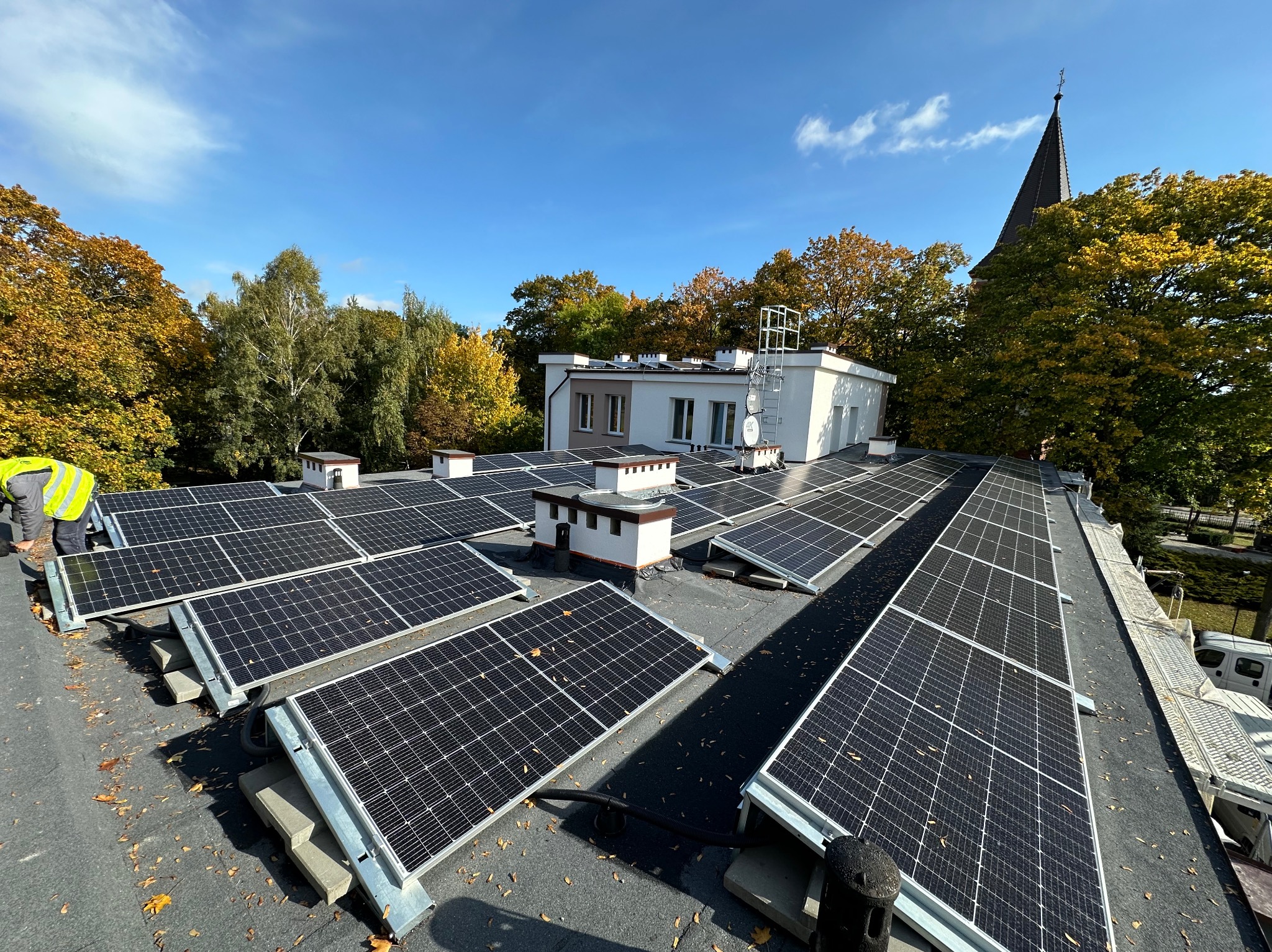 Instalacja paneli fotowoltaicznych na dachu budynku z widoczną wieżą kościoła w tle, jesienne drzewa i błękitne niebo. Częściowo widoczna osoba w odblaskowej kamizelce.