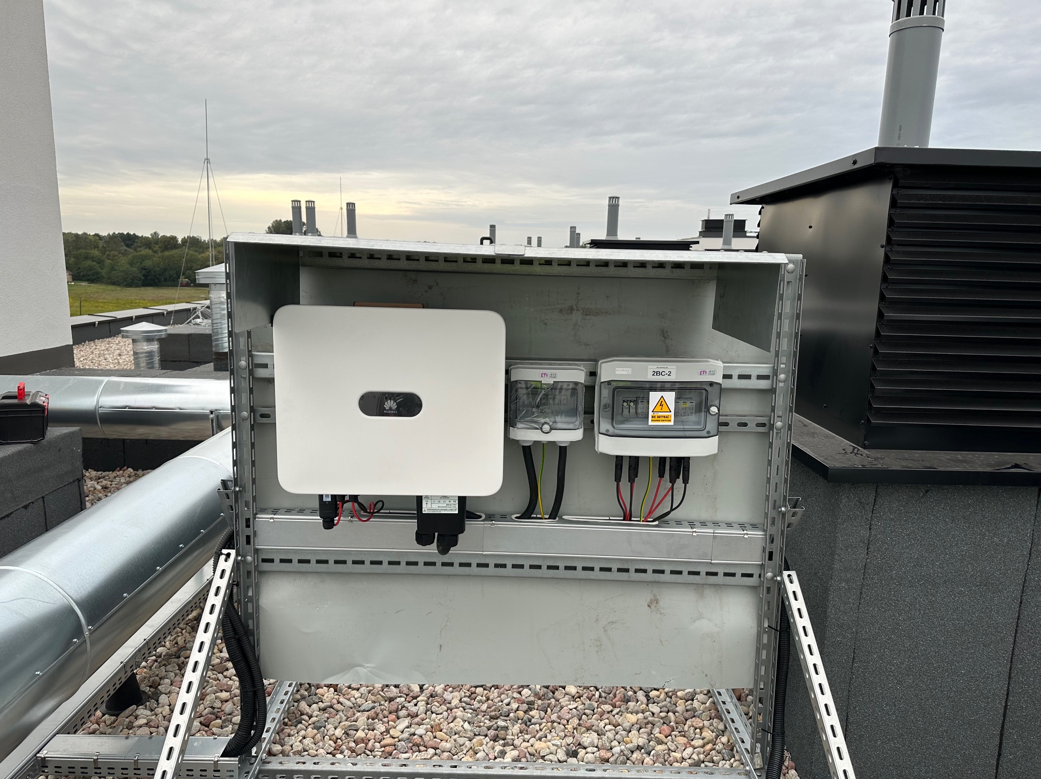 Instalacja fotowoltaiczna na dachu z widocznym inwerterem Huawei i zabezpieczeniami w szarej obudowie. Widoczne przewody i elementy montażowe na tle żwirowego dachu.