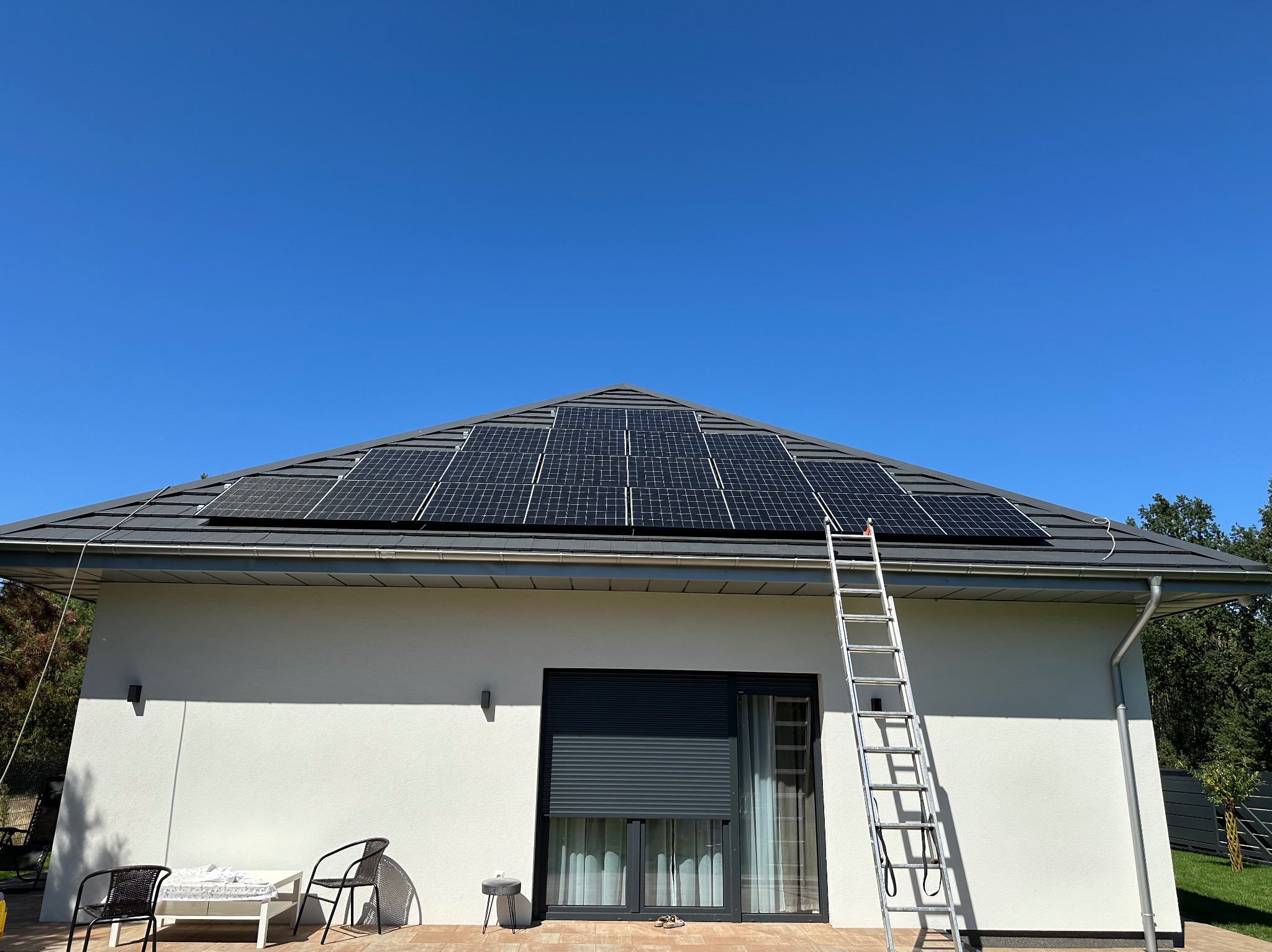 Panele fotowoltaiczne na dachu domu jednorodzinnego w słoneczny dzień. Aluminiowa drabina oparta o dach. Widok na białą elewację i okno z roletą.