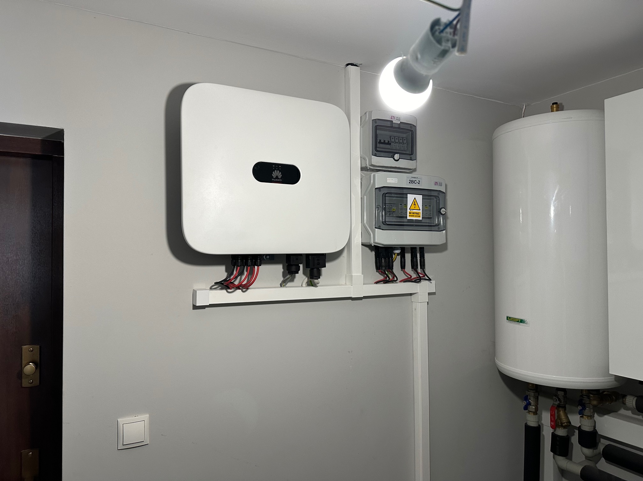 Biały inwerter Huawei i skrzynki elektryczne zamontowane na ścianie wewnątrz pomieszczenia, widoczne przewody i rury. Całość oświetlona żarówką.