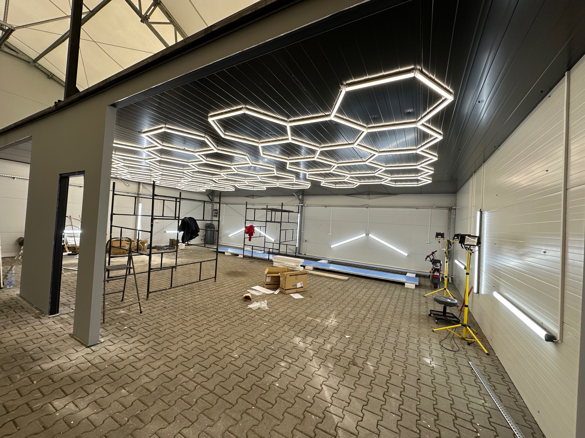 Nowoczesne oświetlenie LED w kształcie plastra miodu na suficie w przestronnym pomieszczeniu. Widoczne metalowe konstrukcje i narzędzia, sugerujące etap montażu.