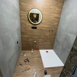 Budownictwo i Usługi - Montaż kabiny prysznicowej w łazience z drewnopodobnymi i szarymi płytkami. Widoczne narzędzia, poziomica laserowa i okrągłe lustro z podświetleniem LED.