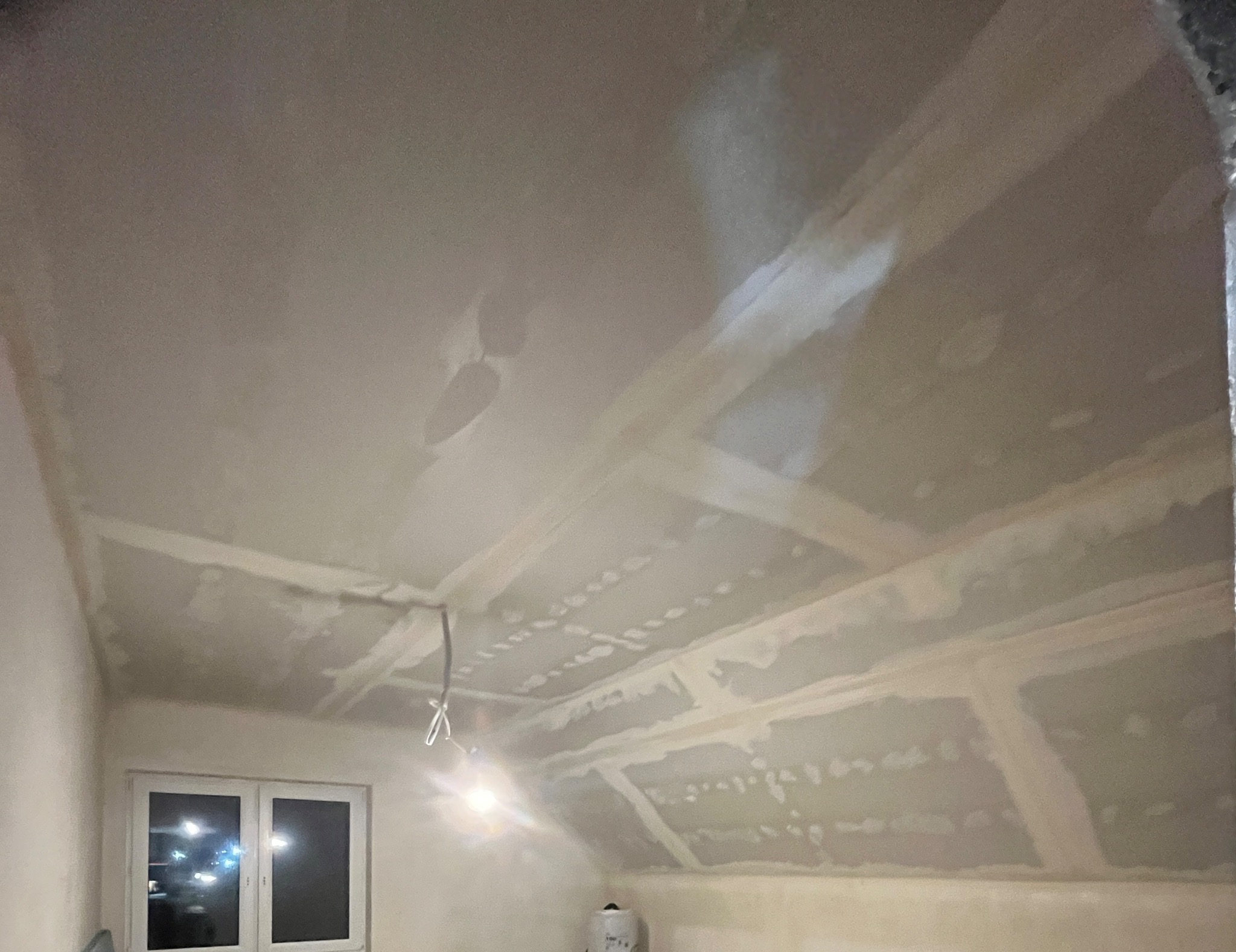 Skośny sufit poddasza po szpachlowaniu, widoczne łączenia płyt gipsowych. Kabel zwisa z sufitu, okno w tle. Praca w toku, przygotowanie do malowania.