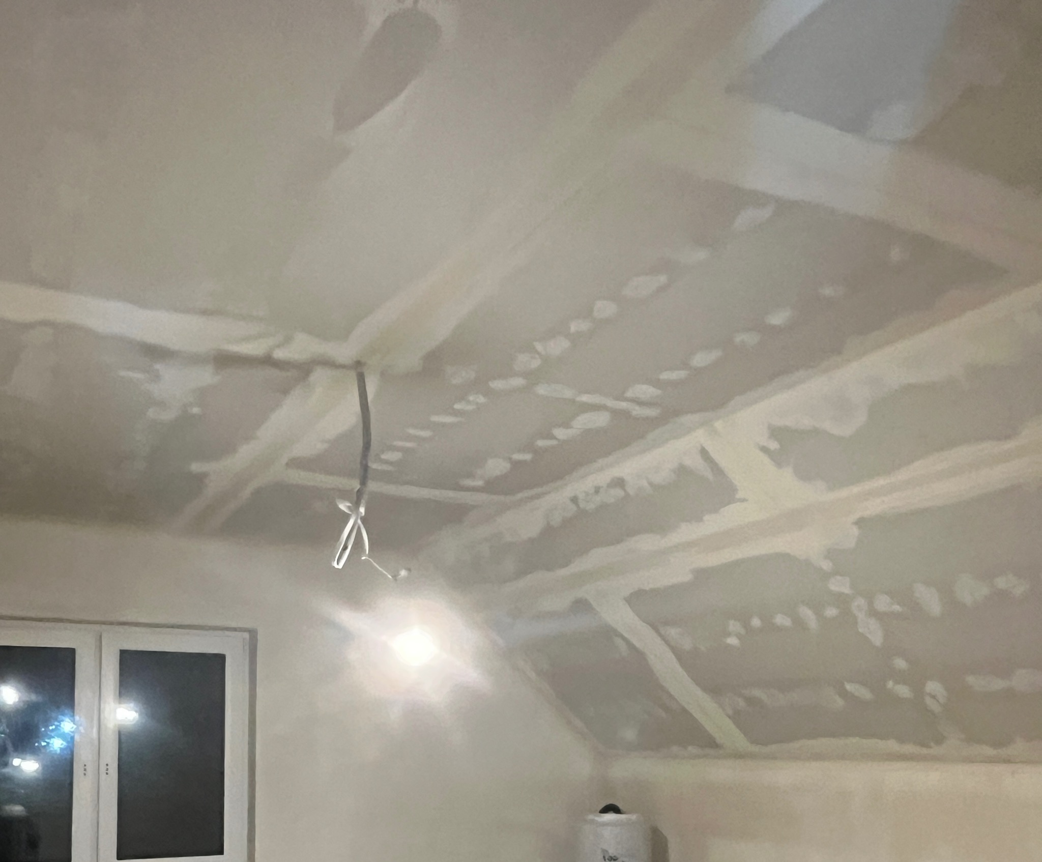 Skośny sufit poddasza po szpachlowaniu, widoczne łączenia płyt gipsowych i wystające kable oświetleniowe. W tle okno i ściana przygotowana do malowania.