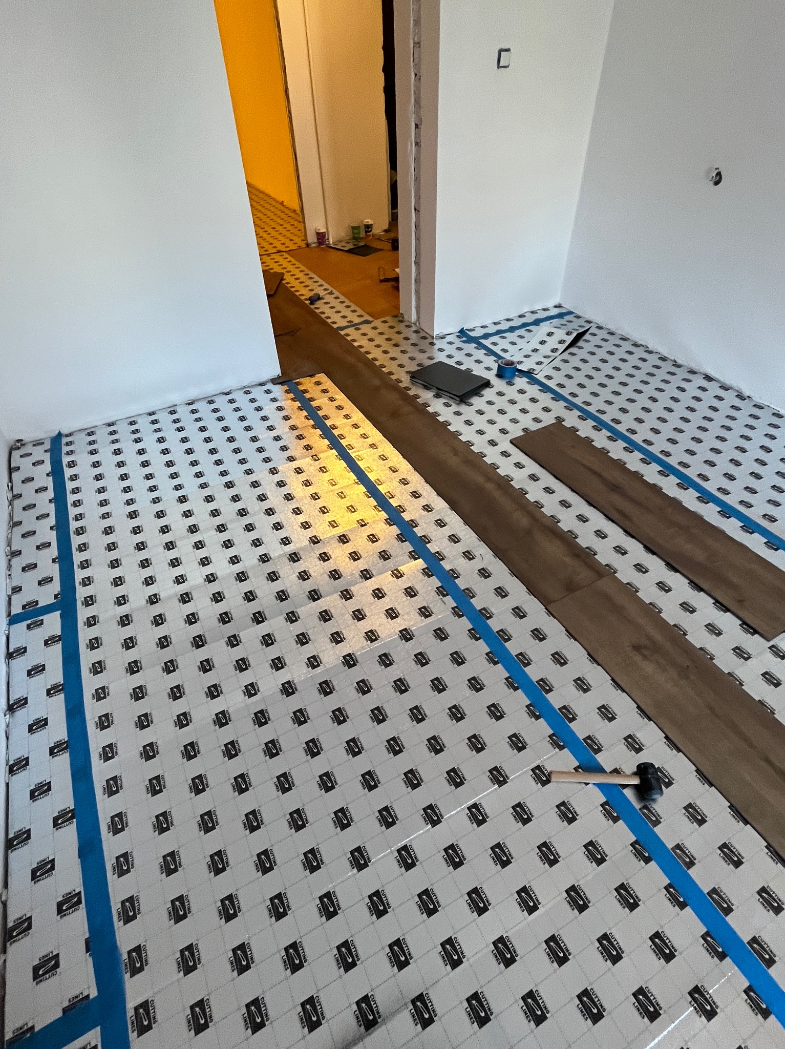 Przygotowanie do montażu paneli podłogowych: rozłożony podkład z taśmą, widoczne deski i młotek. Częściowo ułożona podłoga w tle. Jasne wnętrze, etap prac.