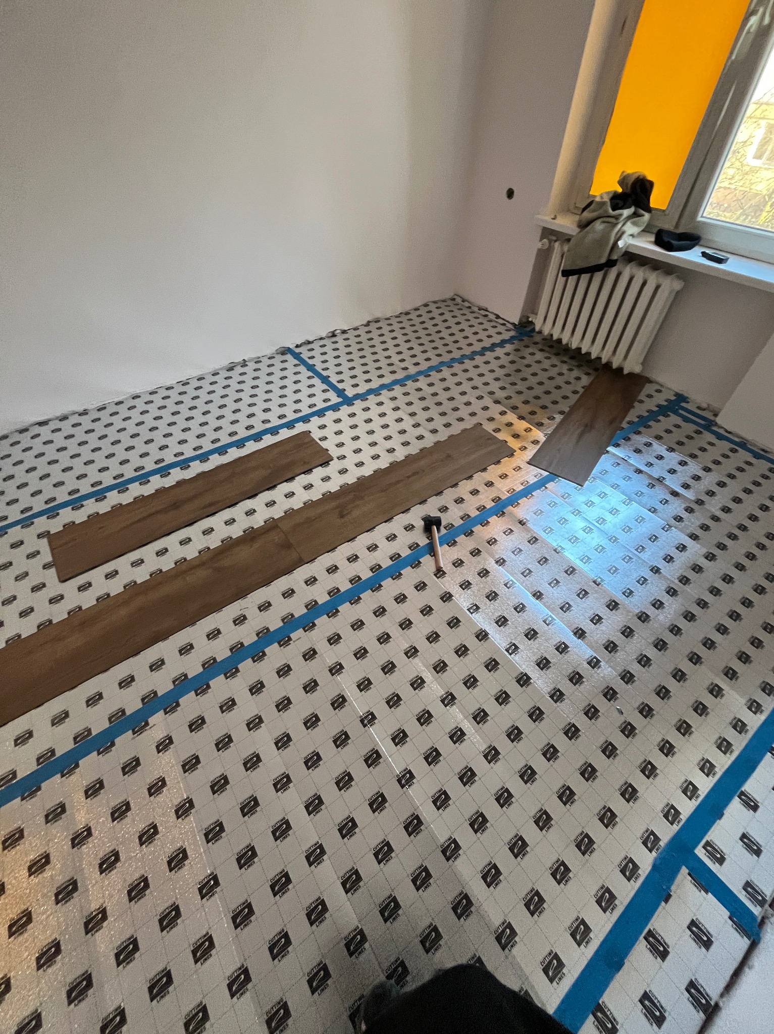 Układanie paneli podłogowych na folii izolacyjnej z użyciem młotka. Widoczne fragmenty paneli i niebieska taśma wyznaczająca obszar roboczy. Przy oknie grzejnik.