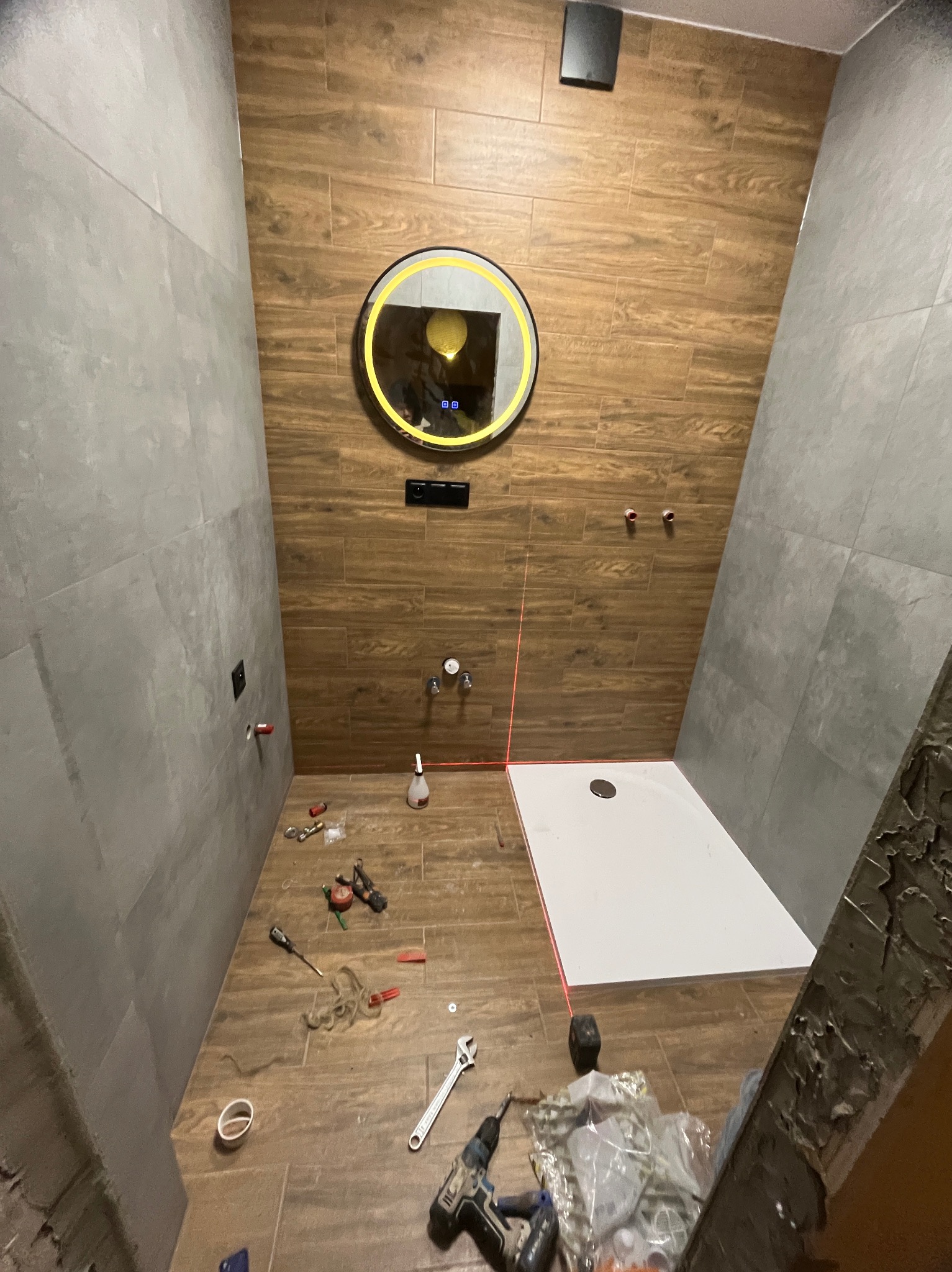 Remont łazienki w trakcie: ułożone płytki imitujące drewno i szare, zamontowany brodzik, poziomica laserowa, narzędzia glazurnicze na podłodze. Okrągłe lustro z podświetleniem.