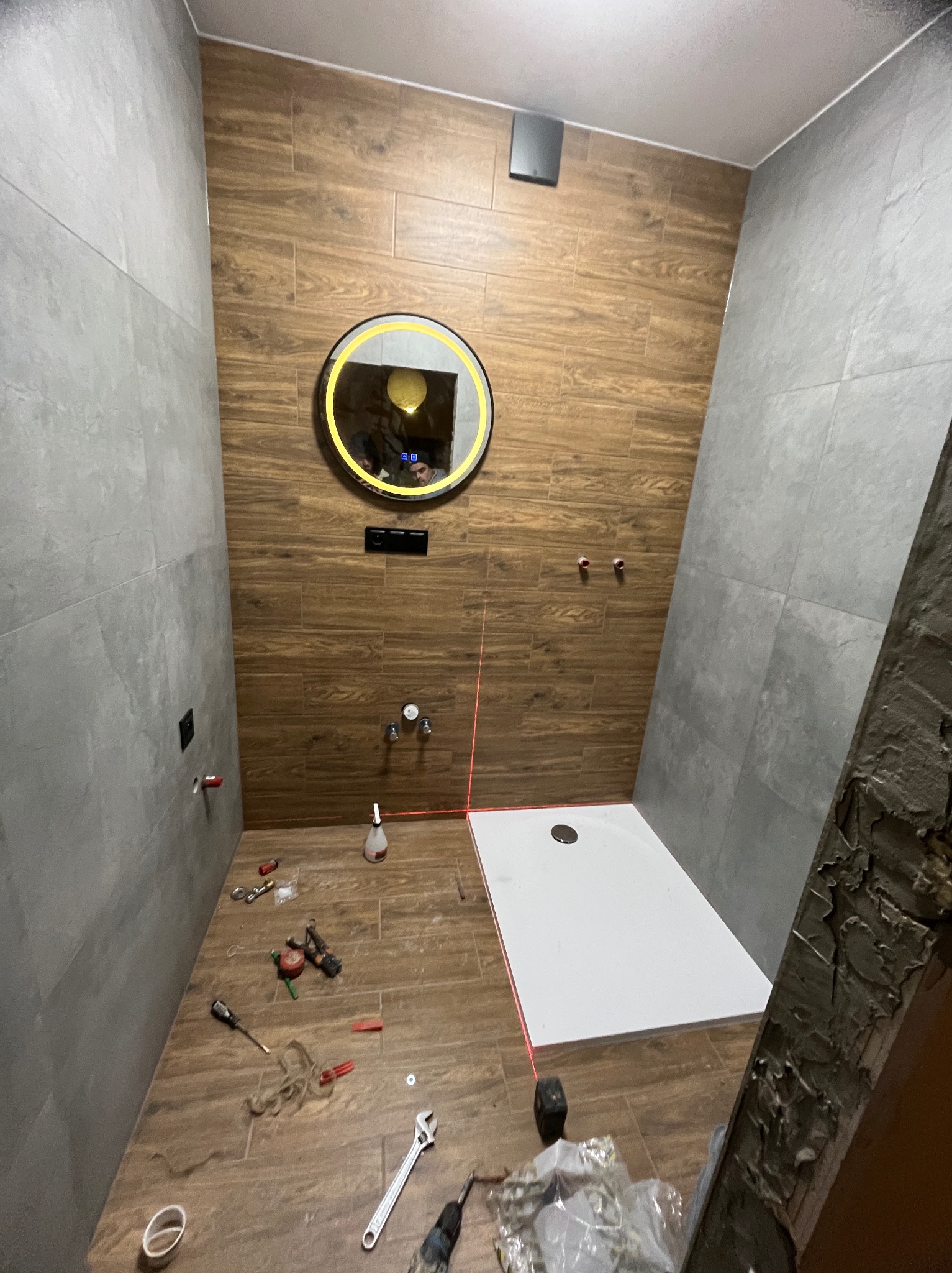 Łazienka w trakcie prac: szare płytki, drewnopodobne panele ścienne, okrągłe lustro z podświetleniem, biały brodzik i narzędzia na podłodze. Widoczny laser krzyżowy.