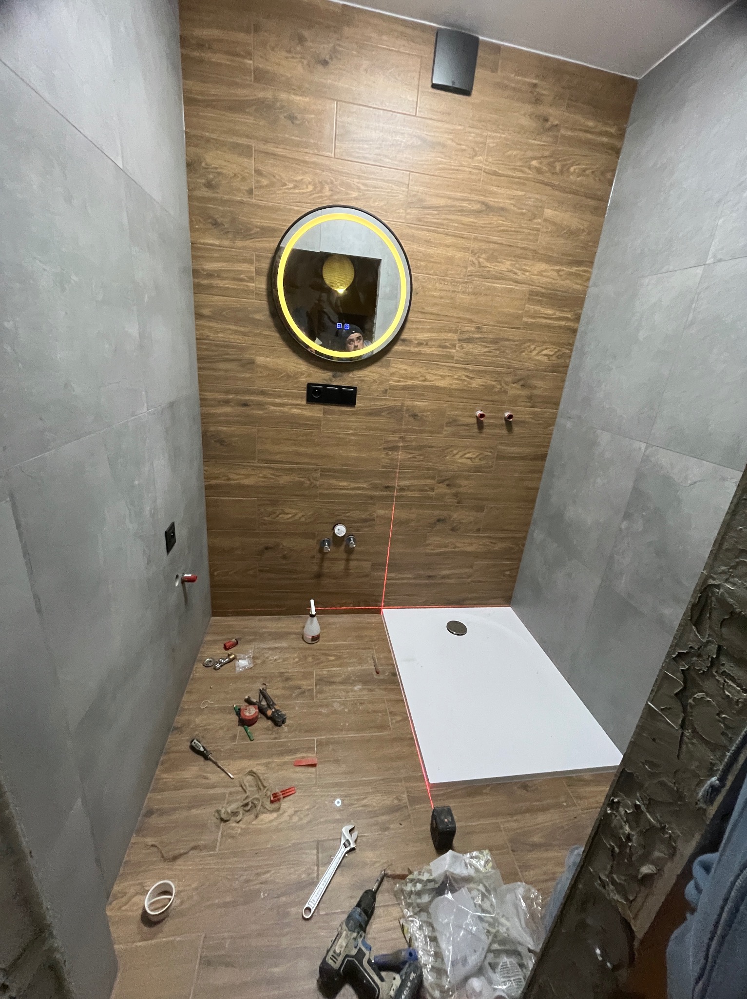 Montaż kabiny prysznicowej w łazience z drewnopodobnymi i szarymi płytkami. Widoczne narzędzia, poziomica laserowa i okrągłe lustro z podświetleniem LED.