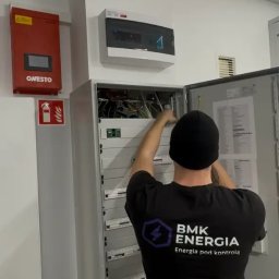 BMK ENERGIA SPÓŁKA Z OGRANICZONĄ ODPOWIEDZIALNOŚCIĄ - Elektryk w czarnej koszulce z logo BMK Energia pracuje przy otwartej szafie elektrycznej w pomieszczeniu z białą ścianą, na której widoczne są urządzenia elektryczne.