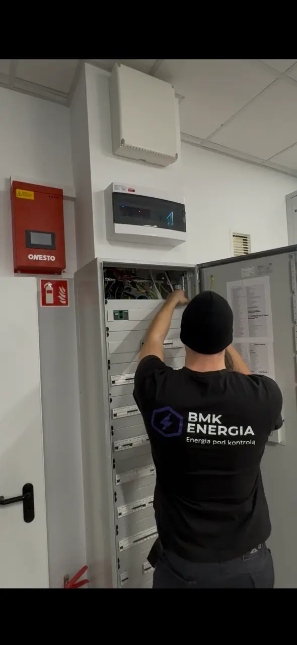 Elektryk w czarnej koszulce z logo BMK Energia pracuje przy otwartej szafie elektrycznej w pomieszczeniu z białą ścianą, na której widoczne są urządzenia elektryczne.