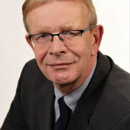 Portret mężczyzny w garniturze, z okularami, uśmiechającego się do kamery na jasnym tle.