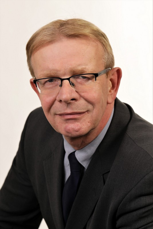 Portret mężczyzny w garniturze, z okularami, uśmiechającego się do kamery na jasnym tle.