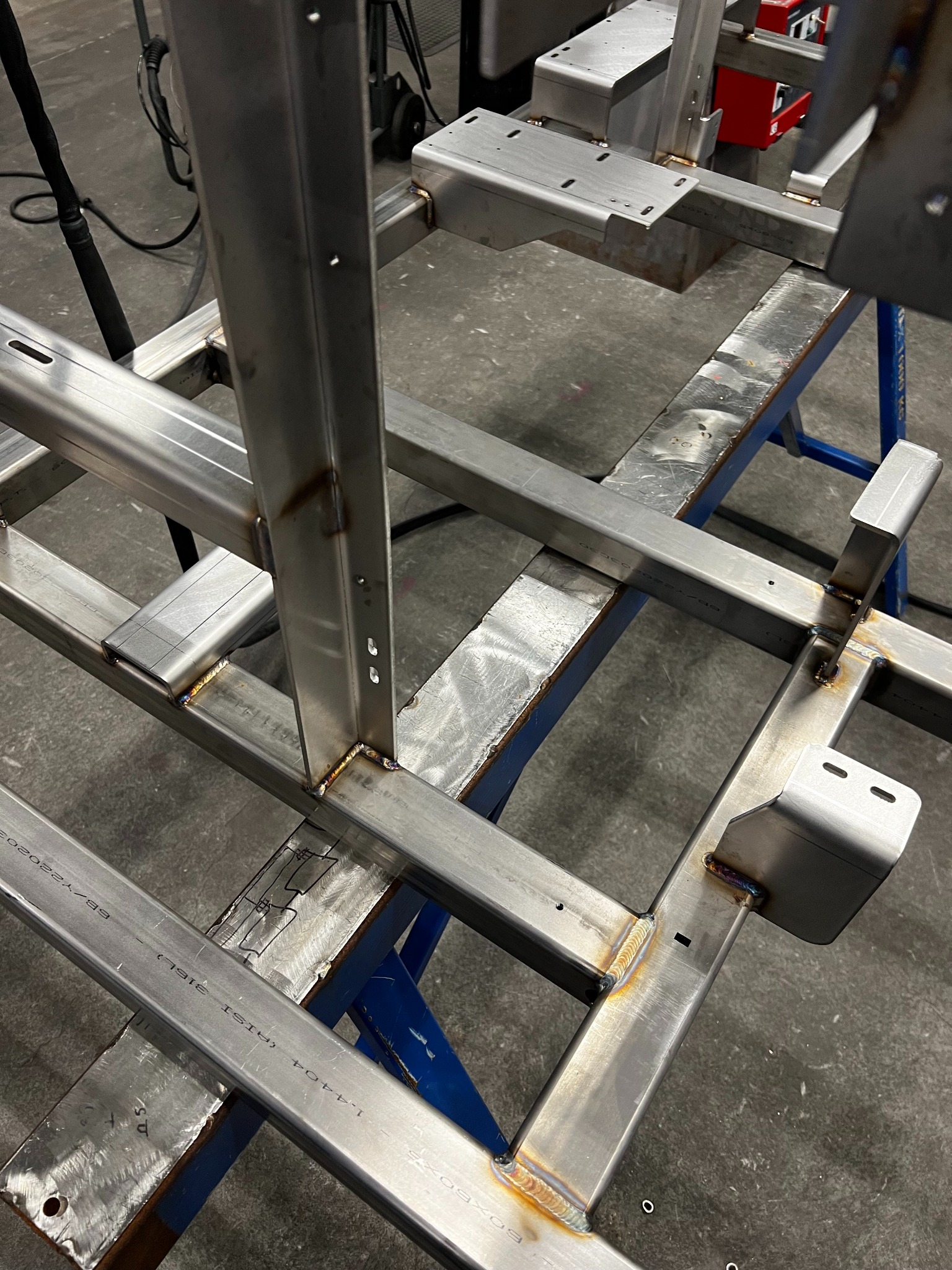 Konstrukcja z profili stalowych po spawaniu, widoczne spoiny, otwory i oznaczenia na metalu. Elementy wsparte na niebieskim stojaku warsztatowym.