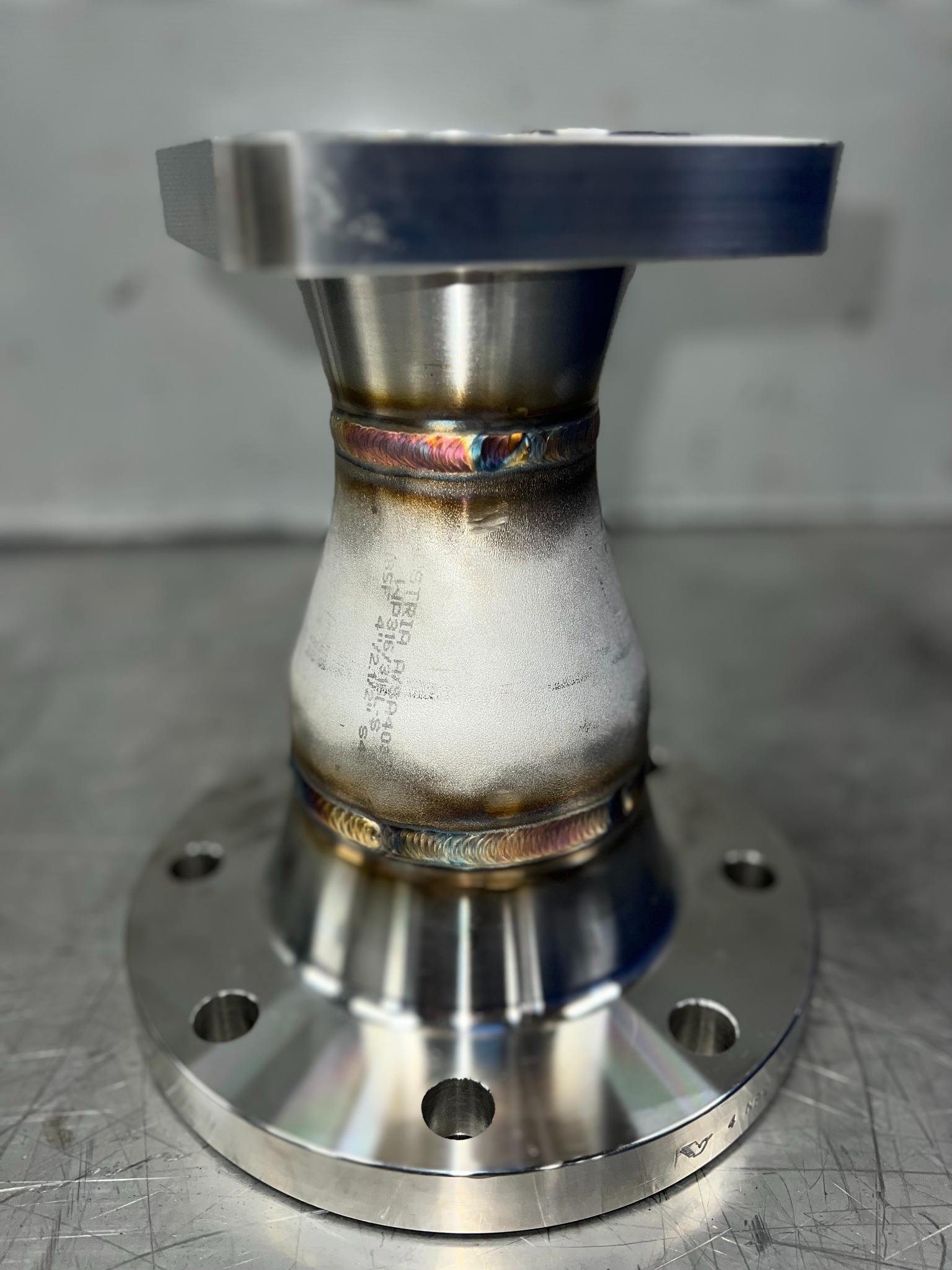 Precyzyjnie spawany element metalowy z widocznymi śladami spawania i oznaczeniami, umieszczony na metalowej powierzchni. Detal z bliska, ukazujący jakość wykonania.