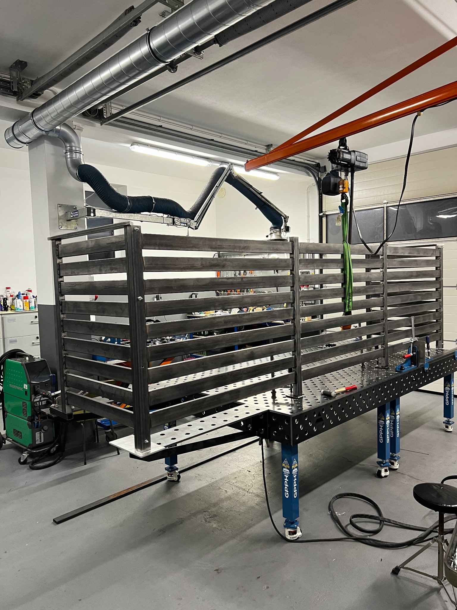 Spawana konstrukcja metalowa z poziomymi szczeblami na stole spawalniczym w warsztacie w Łomazach. Widoczny odciąg spalin i spawarka Migatronic.