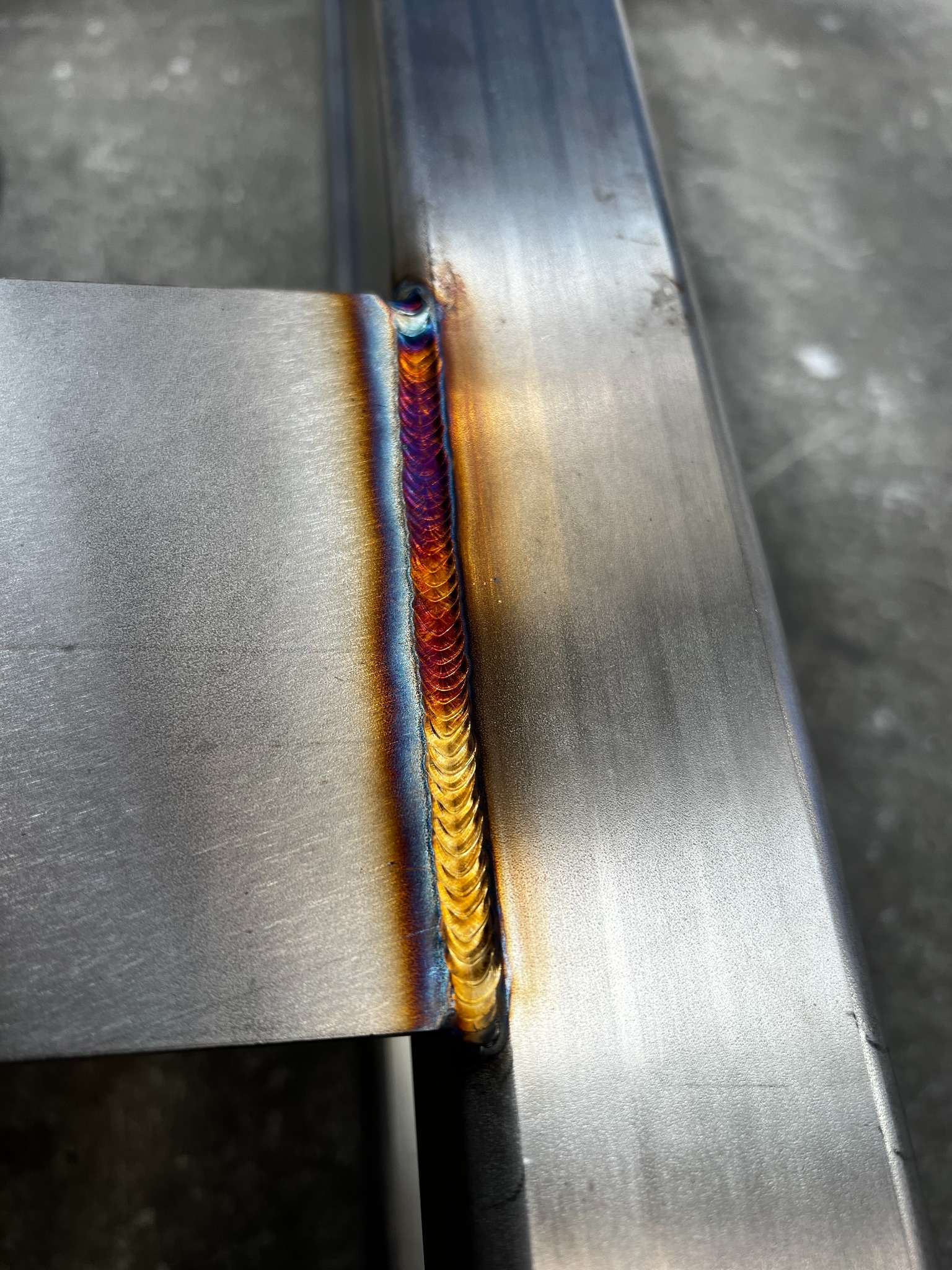 Metalowa konstrukcja po spawaniu, widoczny precyzyjny spaw z charakterystycznymi kolorami przegrzanego metalu. Detal z bliska, ukazujący jakość wykonania.