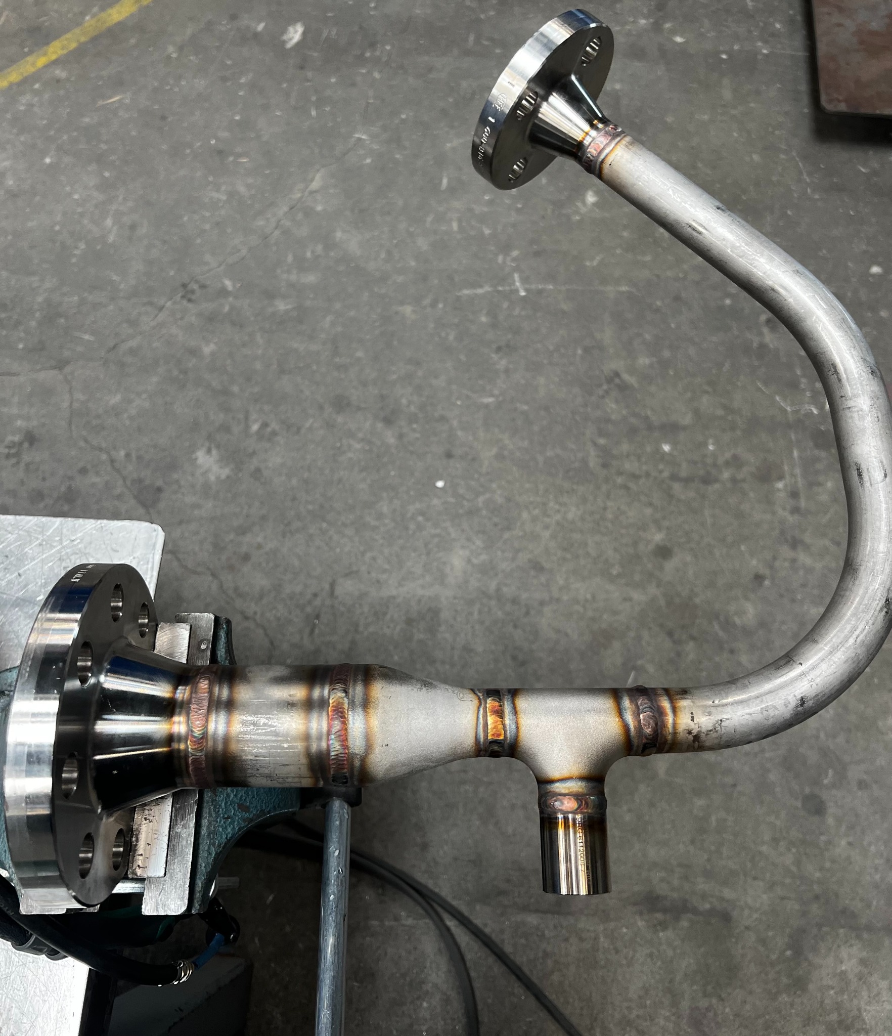 Precyzyjnie spawana, nierdzewna konstrukcja rurowa z kołnierzami, zamocowana w imadle. Widoczne spoiny i szlifowane powierzchnie. Detal obróbki metalu.