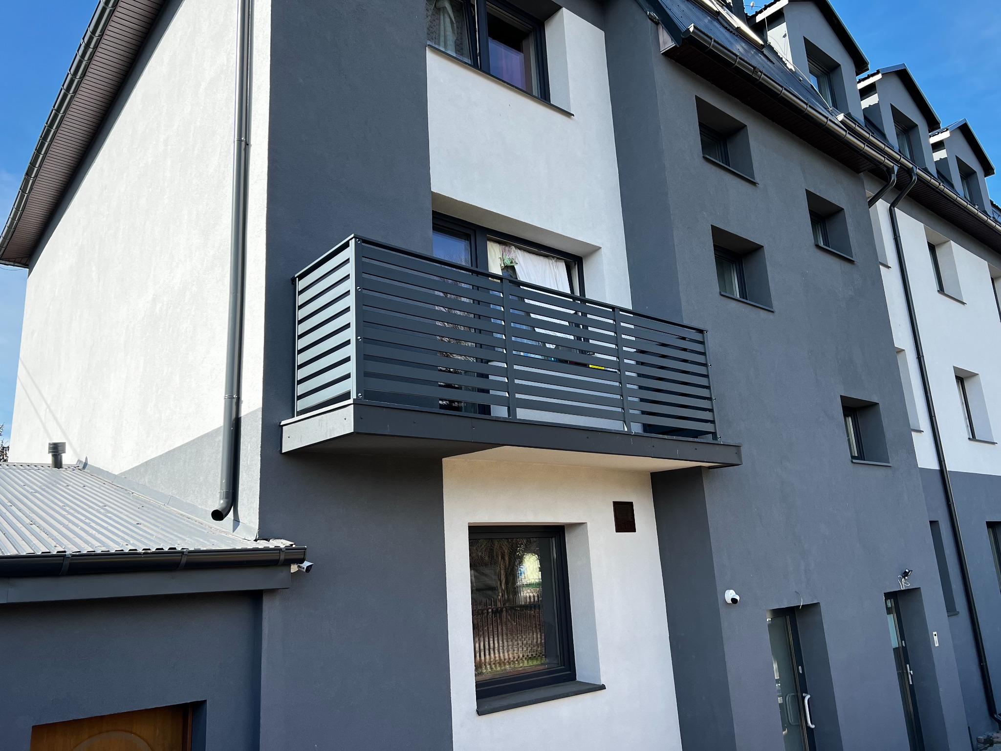 Nowoczesny balkon z poziomą, grafitową konstrukcją, wkomponowany w elewację budynku o szaro-białej kolorystyce. Widok z dołu, podkreślający architekturę.