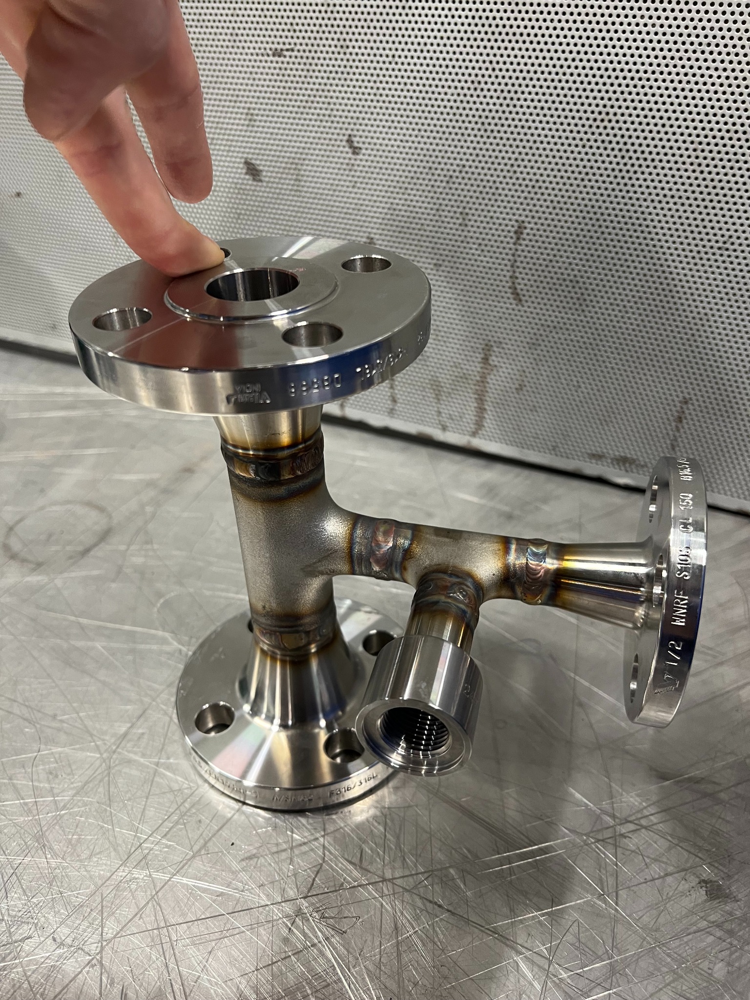 Precyzyjnie spawany, trójnik ze stali nierdzewnej z kołnierzami, prezentowany na metalowym stole. Palec wskazujący na górny kołnierz. Widoczne spawy i oznaczenia.