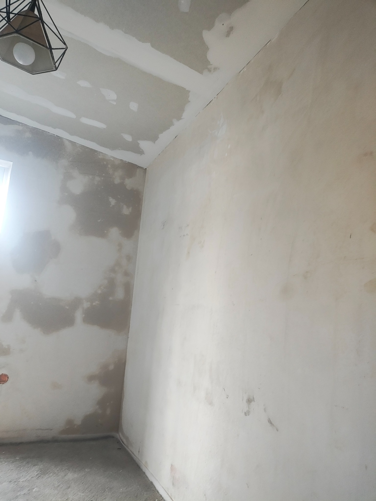 Pokój w trakcie szpachlowania ścian i sufitu, widoczne nierówności i miejsca po łataniu. W rogu sufitu lampa z metalowym kloszem.