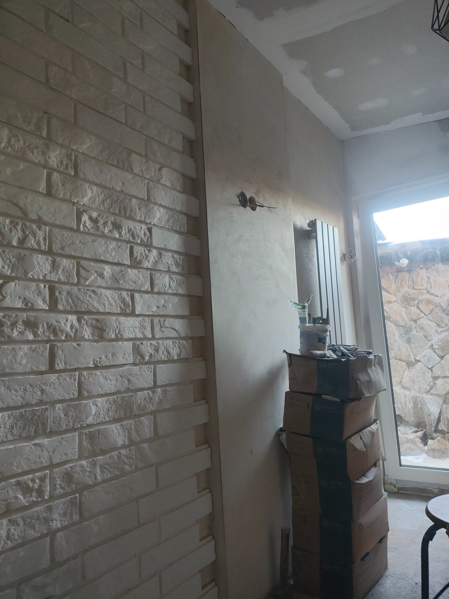 Wykończona ściana z dekoracyjnej białej cegły w pomieszczeniu w trakcie remontu. Widoczne kartony z materiałami budowlanymi i grzejnik.