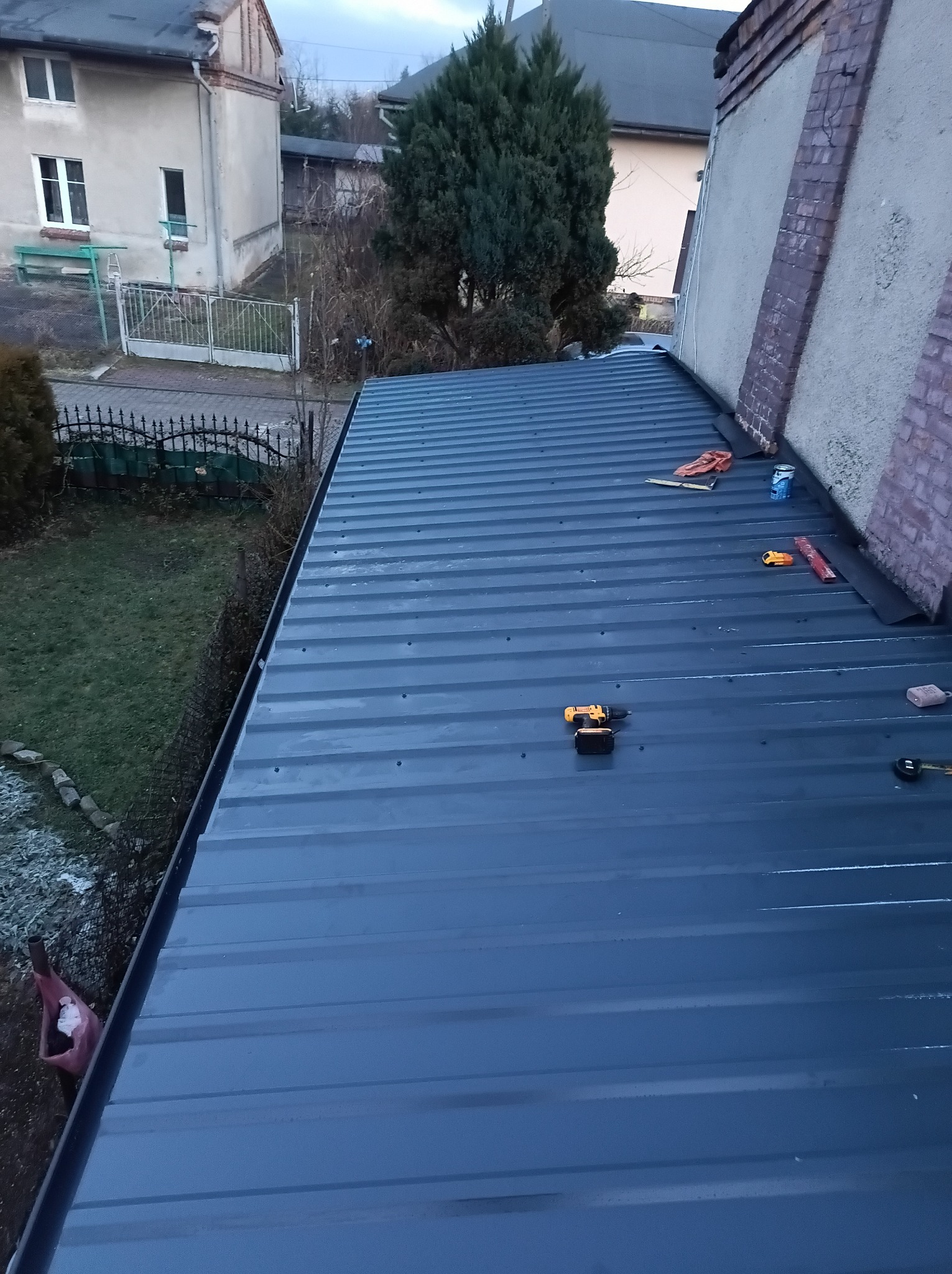 Nowy, szary dach z blachy trapezowej, widok z góry. Na dachu leżą narzędzia: wkrętarka, miarka, puszka farby. W tle budynki mieszkalne i zieleń.