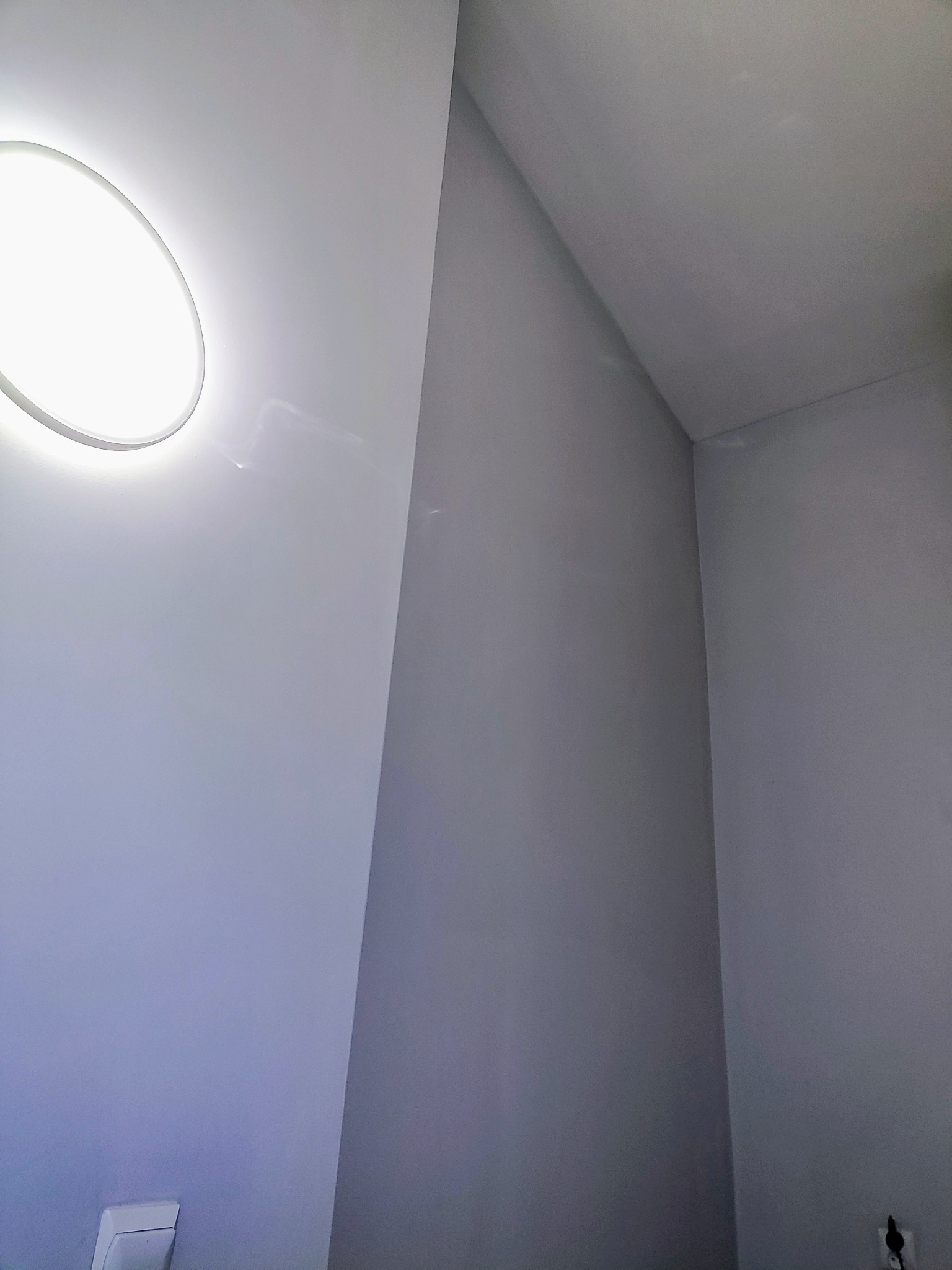 Świeżo pomalowane, gładkie ściany w odcieniach szarości, oświetlone okrągłą lampą sufitową. Widoczny narożnik i gniazdko elektryczne.