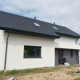 MR BUD - Nowoczesny dom jednorodzinny z białą elewacją i grafitowym dachem. Widoczny fragment elewacji w trakcie prac wykończeniowych przy wejściu.