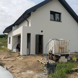 MR BUD - Nowoczesny dom z jasną elewacją i ciemną stolarką okienną. Widoczny pracownik podczas prac wykończeniowych. Na pierwszym planie zbiornik na wodę i materiały budowlane.