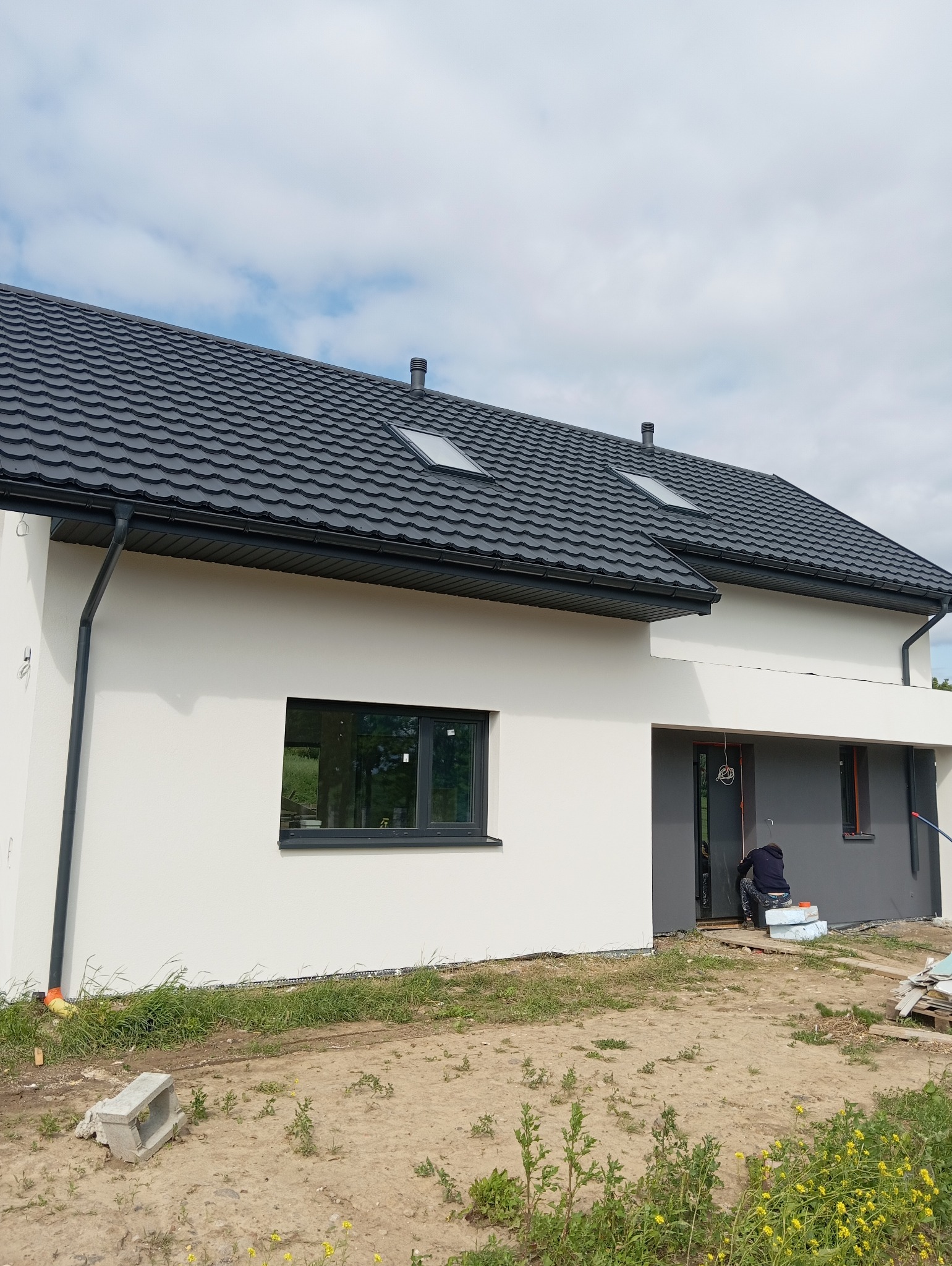 Nowoczesny dom jednorodzinny z białą elewacją i grafitowym dachem. Widoczny fragment elewacji w trakcie prac wykończeniowych przy wejściu.