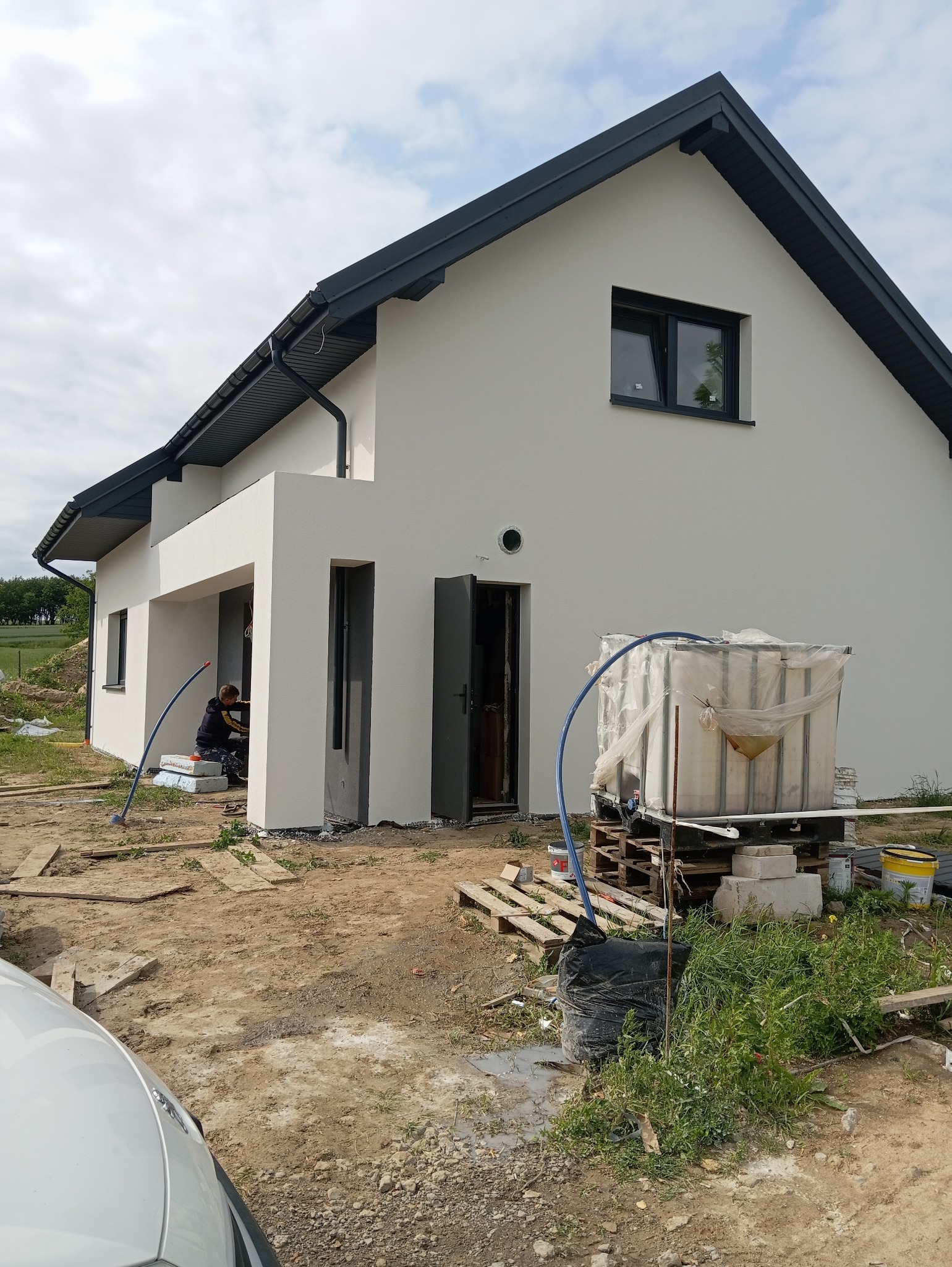 Nowoczesny dom z jasną elewacją i ciemną stolarką okienną. Widoczny pracownik podczas prac wykończeniowych. Na pierwszym planie zbiornik na wodę i materiały budowlane.