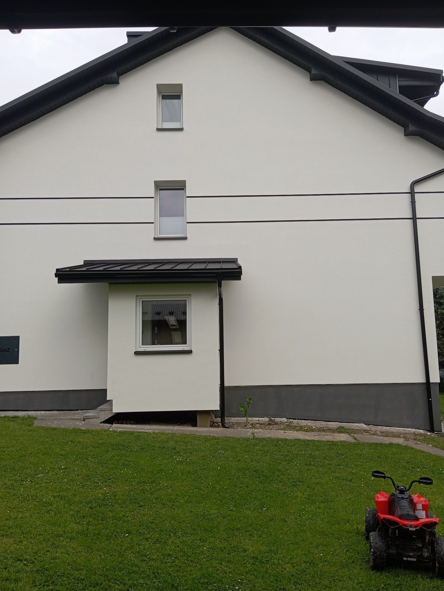 Biała elewacja domu jednorodzinnego z czarnymi akcentami, widok z poziomu trawnika, na którym stoi czerwony quad dla dzieci. Nowoczesny design.