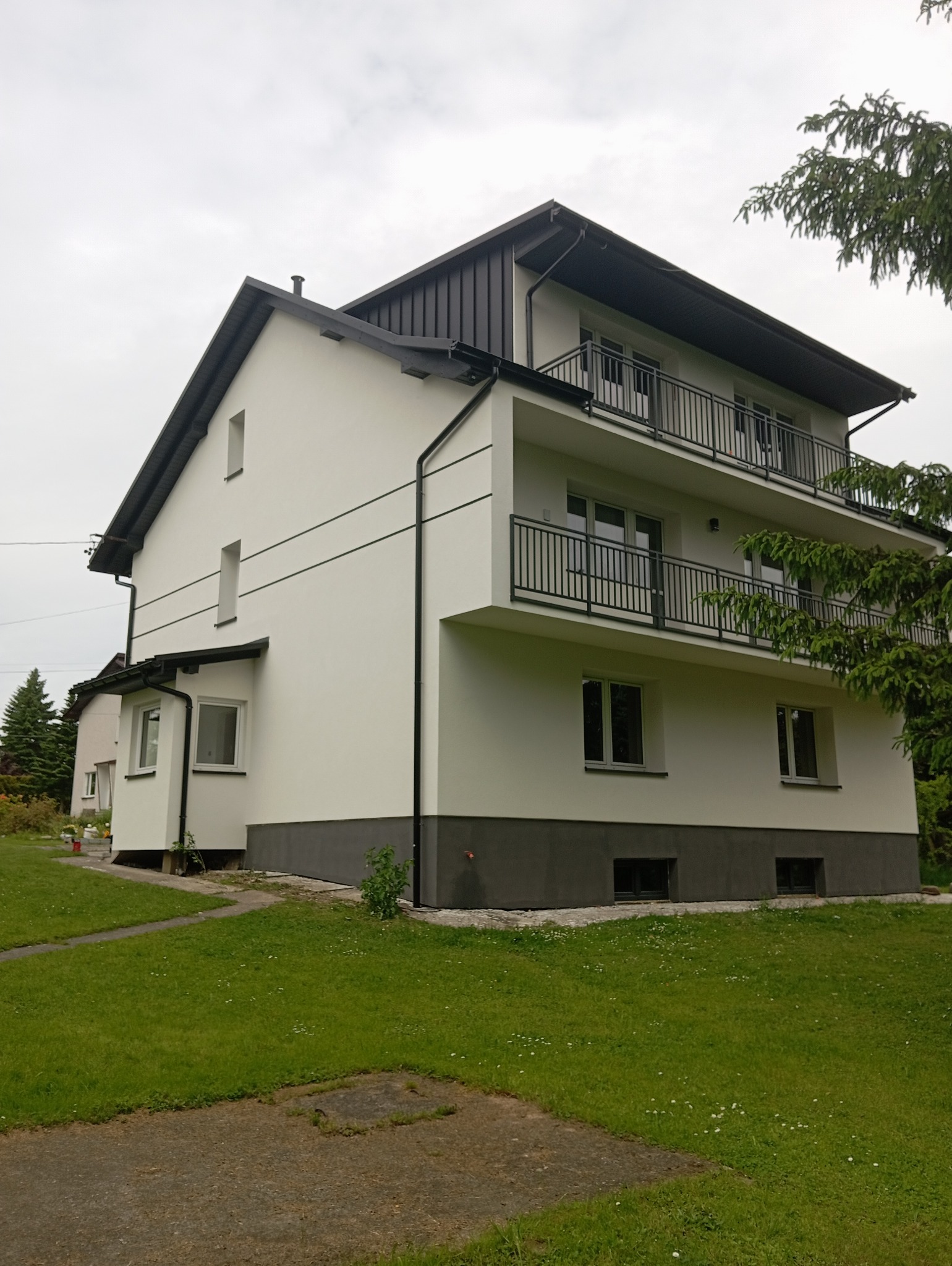 Dwupiętrowy dom z białymi ścianami i ciemnoszarą podmurówką, balkonami z czarną balustradą. Widok z boku na tle zielonego trawnika i pochmurnego nieba.