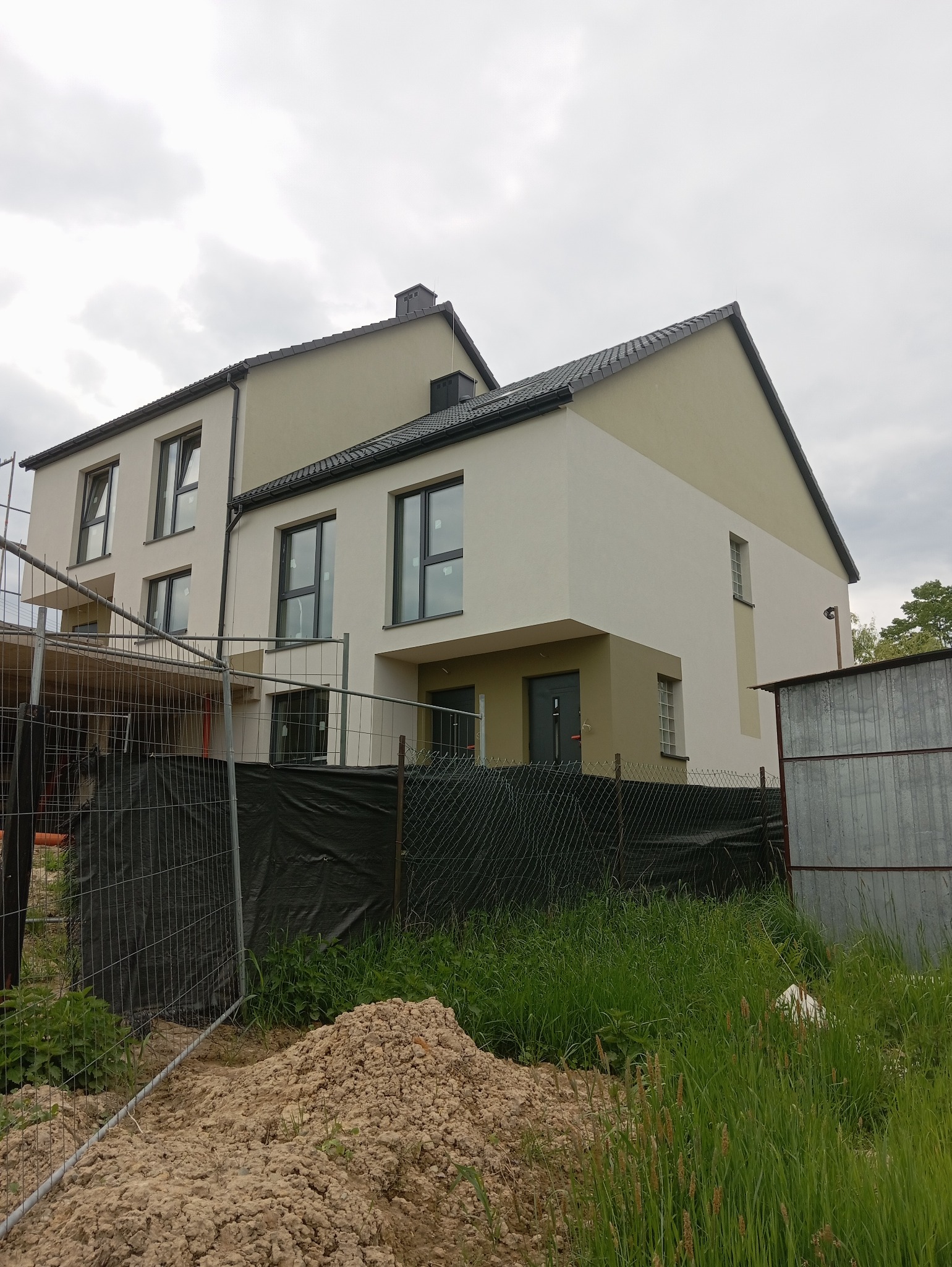 Dwukondygnacyjny dom z płaskim dachem, świeżo po malowaniu elewacji w odcieniach bieli i beżu, widoczny zza ogrodzenia budowy i sterty ziemi.
